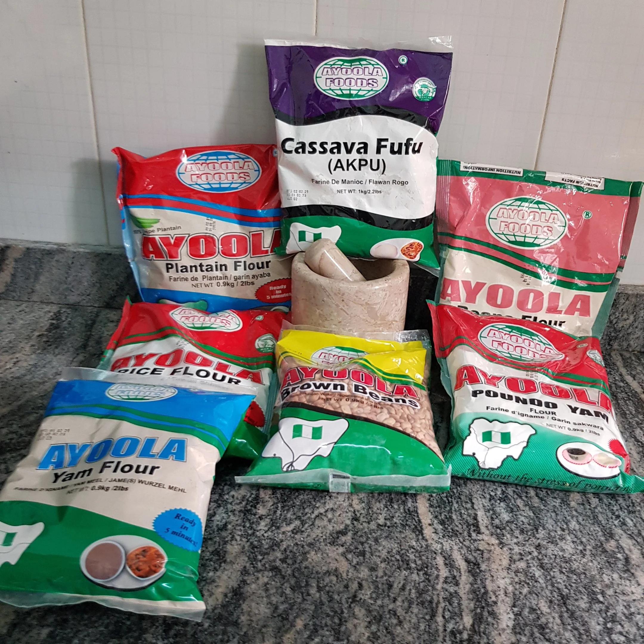 Various bags of Ayoala brand flours, including Plantain Flour, Cassava Fufu, Yam Flour, Brown Beans, Pounded Yam, and Yam Flour, along with a mortar and pestle on a kitchen countertop.