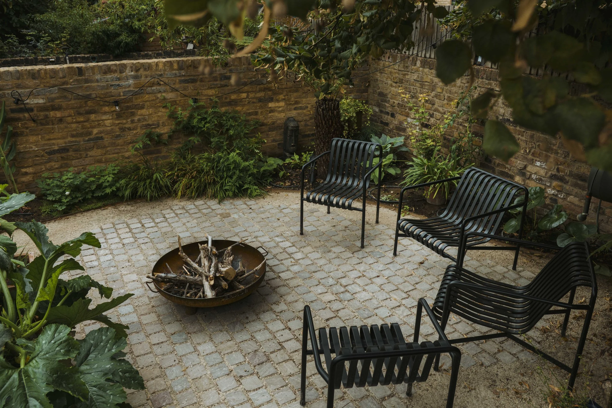 Outdoor patio area with four black metal chairs arranged around a fire pit filled with logs, surrounded by green plants and enclosed by a brick wall.
