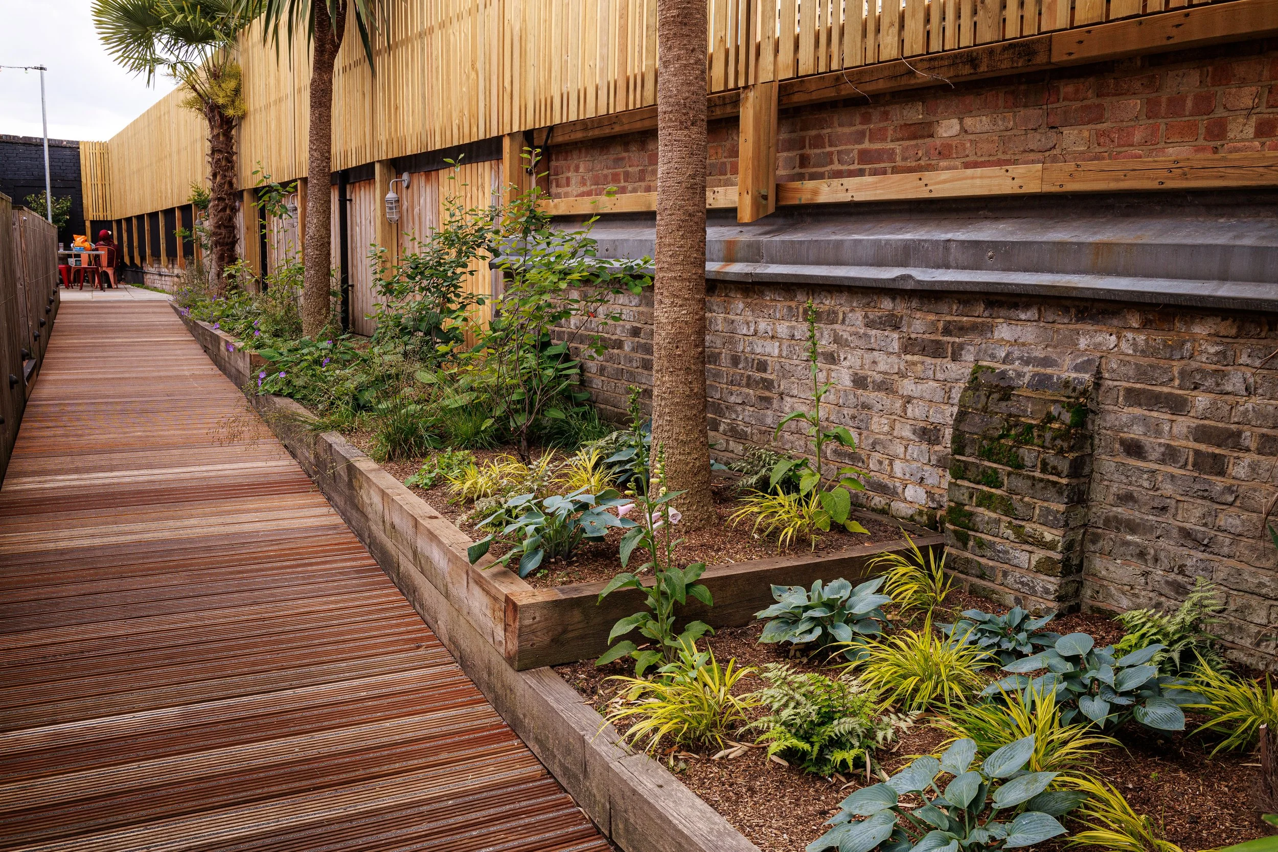 Wooden walkway alongside a garden bed with various plants and trees, bordered by a brick wall and wooden fencing.