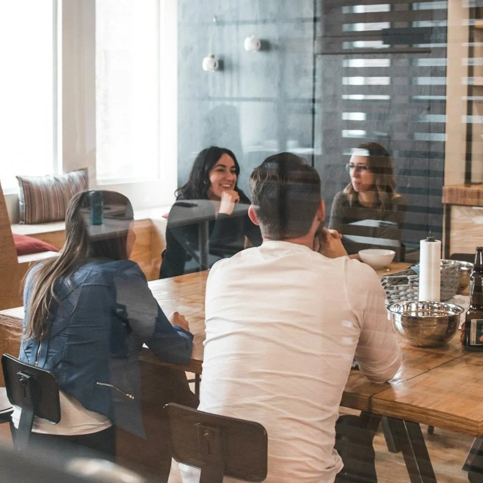 Four people are sat around a table having a discussion, the photo has been taken through a glass partition.