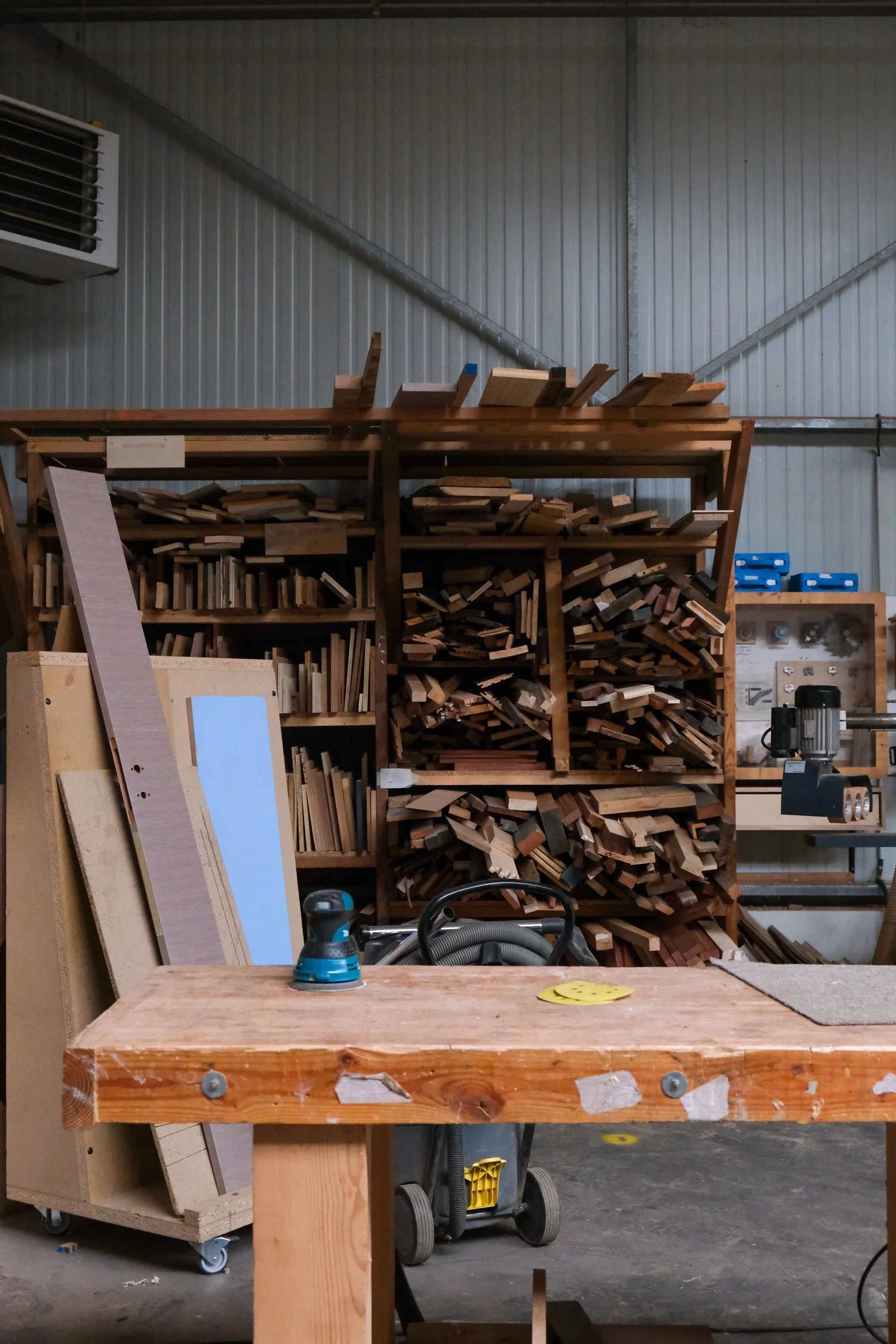 Un atelier de menuiserie avec des étagères remplies de morceaux de bois, une table en bois avec un outil électrique et une planche à découper, murs en métal.