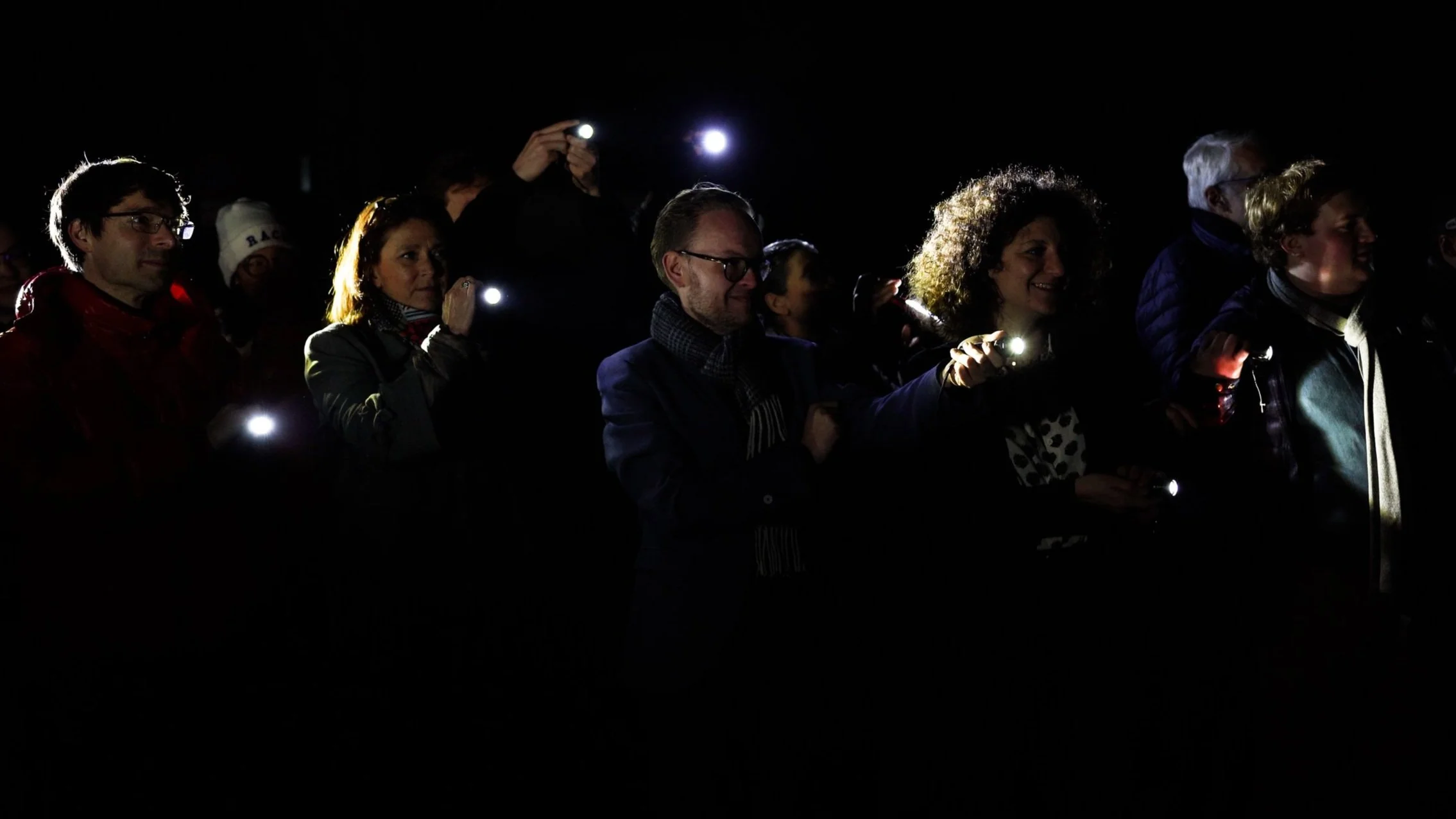 Groupe de personnes dans l'obscurité, tenant des lampes de poche allumées, assistant à un événement nocturne.