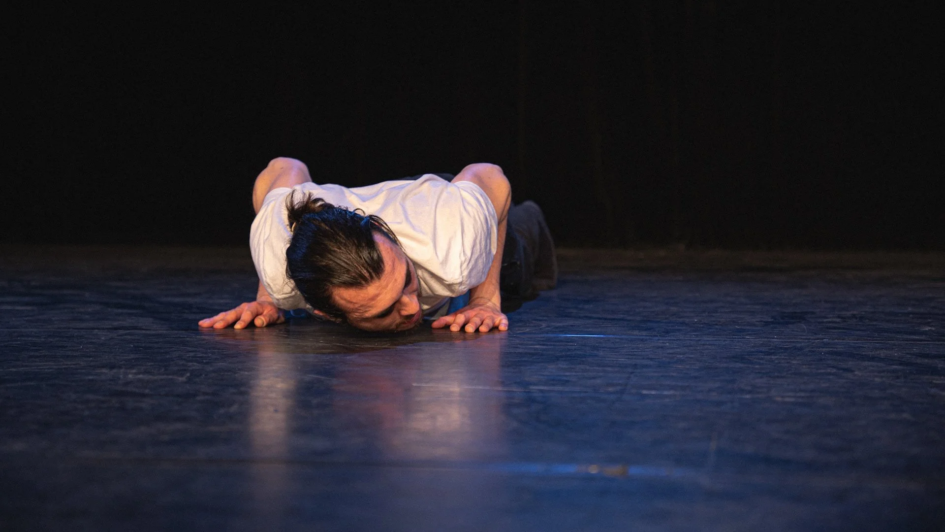 Une personne à genoux sur un sol sombre, appuyée sur les mains et le front, dans une posture expressive ou dramatique.