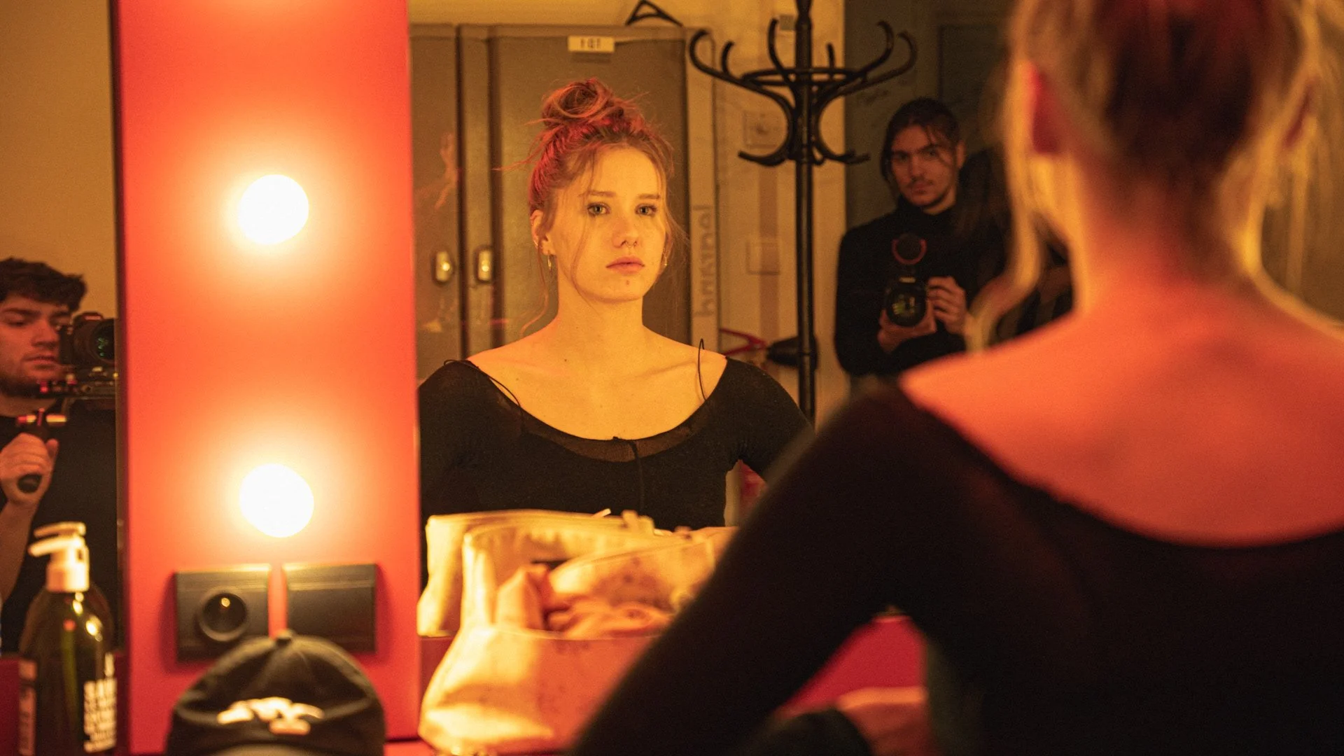 Une femme regarde son reflet dans un miroir derrière une vanité avec des lumières, dans une pièce aux lumières chaudes, avec un homme filmant et un autre photographe en arrière-plan.