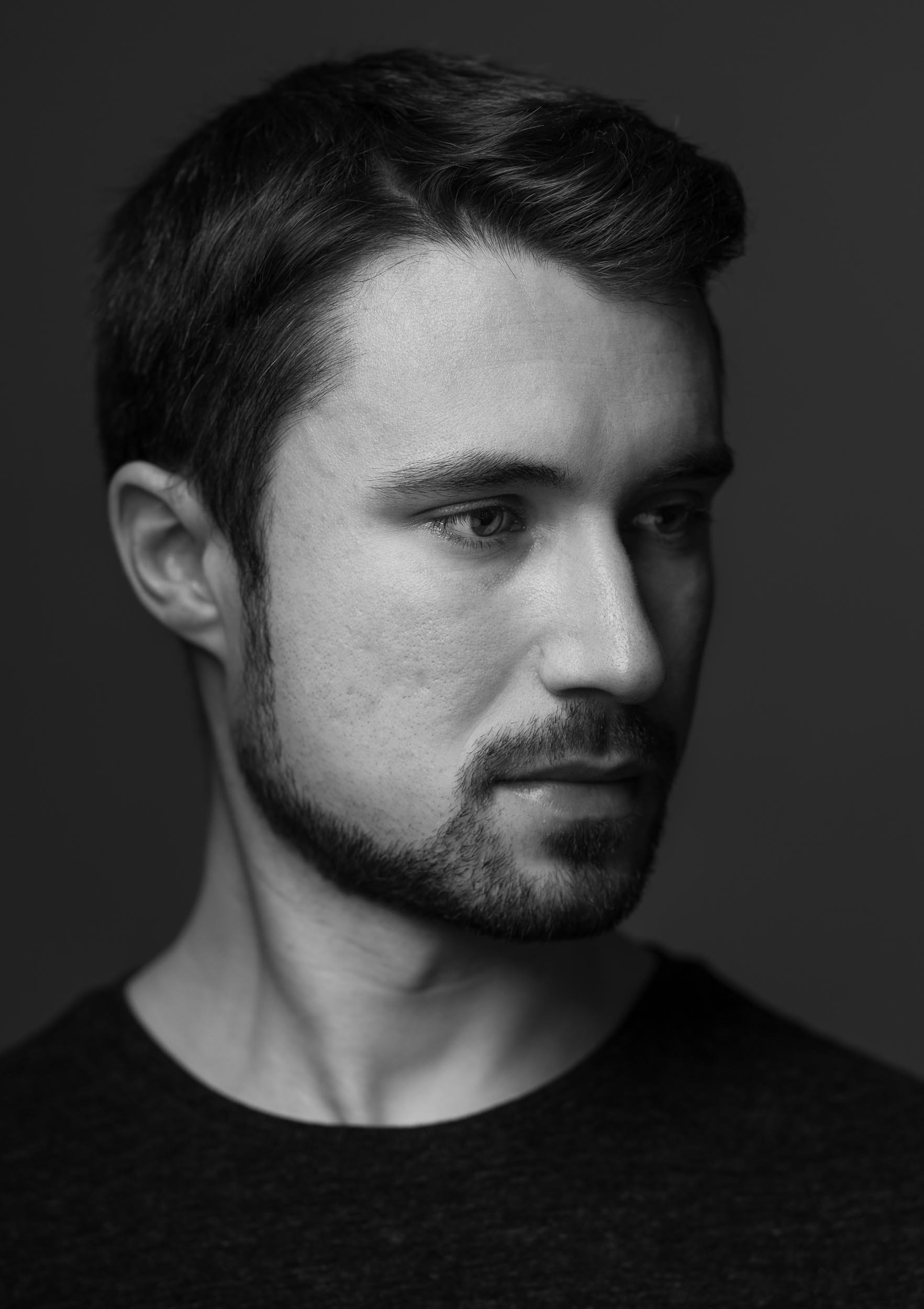 Schwarz-weiß Portrait eines jungen Mannes mit dunklen Haaren und Bart, nachdenklich blickend, dunkler Hintergrund.