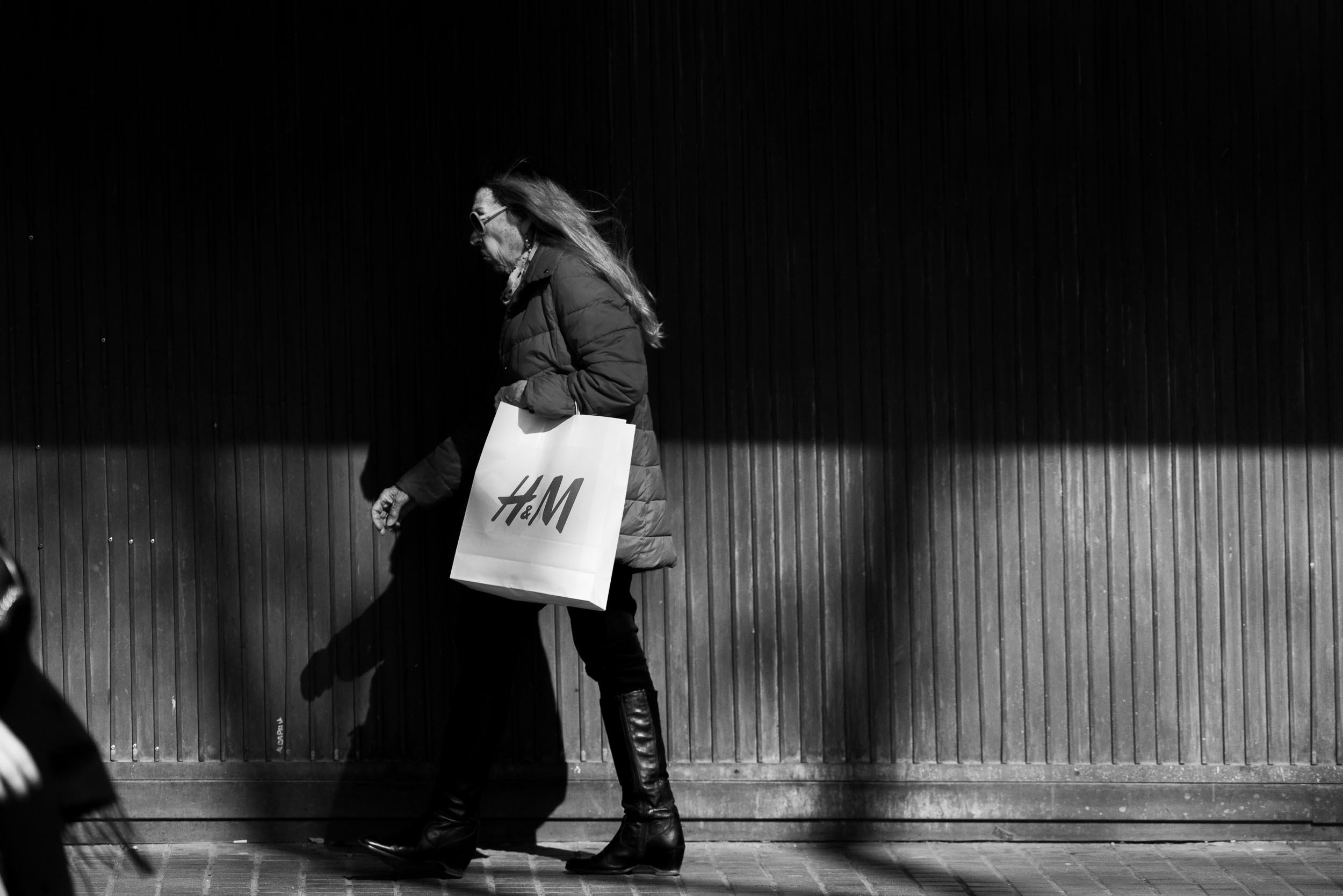 Frau mit langem Haar in Wolkenkratzer-Kleidern und Brille, die einen H&M Einkaufssack trägt, geht an einer Wand vorüber, schwarze und weiße Fotografie.