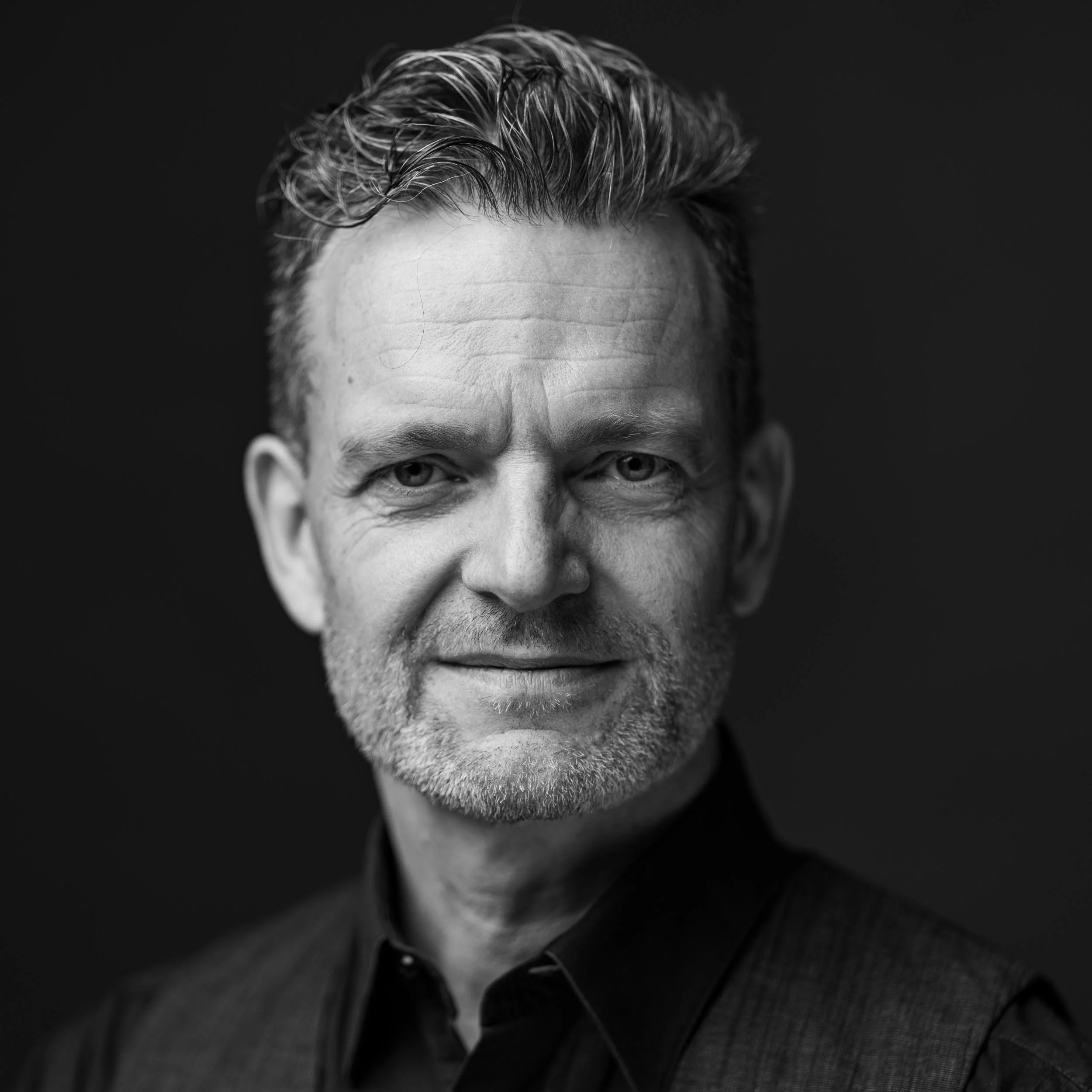 Schwarz-weiß Porträt eines Mannes mit grauem Bart, kurze Haare, trägt einen dunklen Hemd und einen Weste, schaut lächelnd in die Kamera.