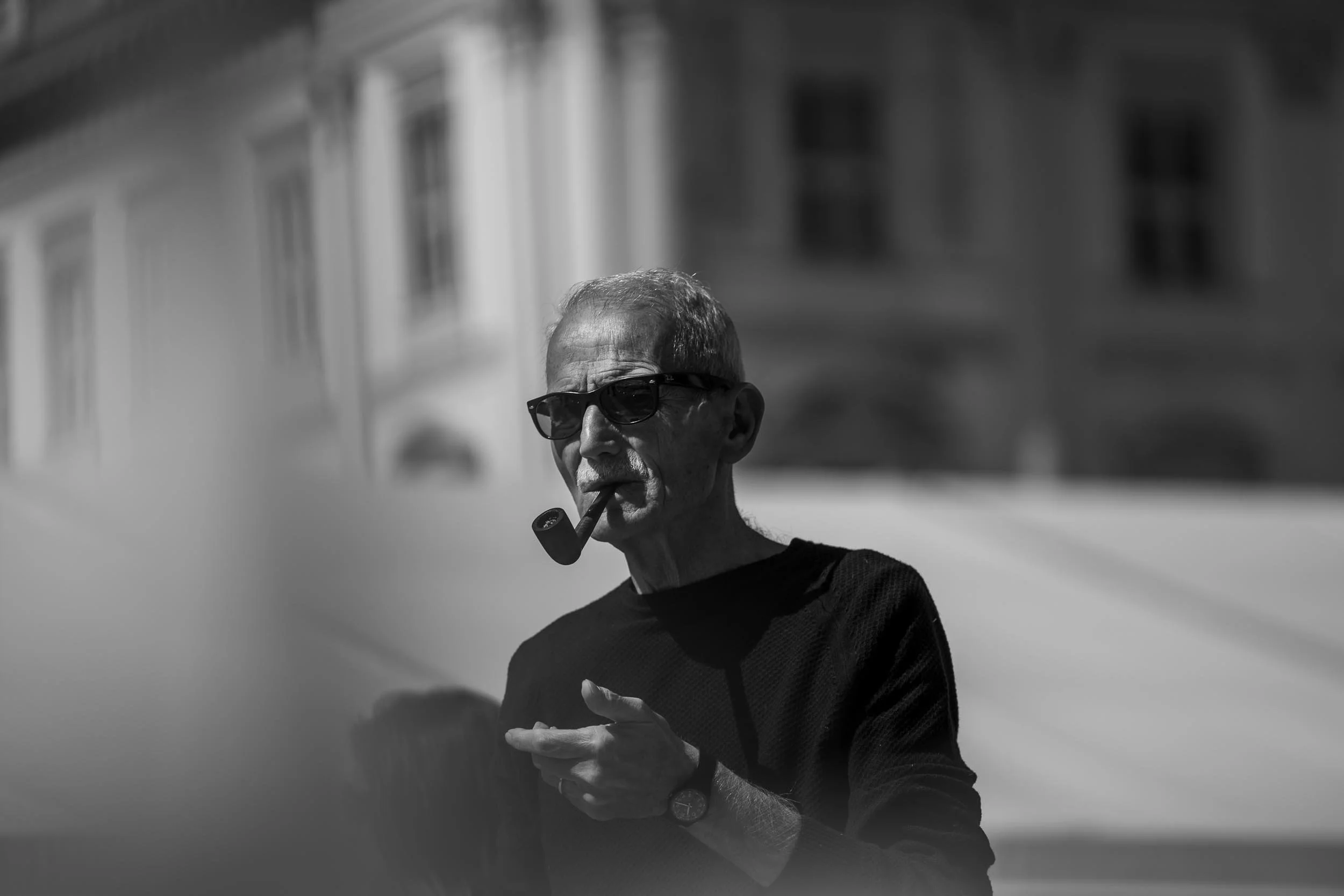 Ein älterer Mann trägt Sonnenbrille, raucht an einer Pfeife und trägt eine Armbanduhr, während er draußen bei sonnigem Wetter steht.