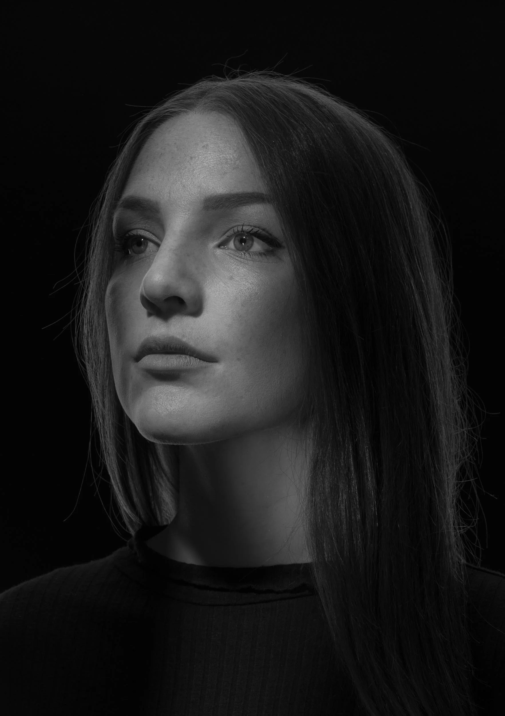 Schwarzweiß-Porträt einer jungen Frau mit langen, glatten Haaren, die nach links schaut, vor dunklem Hintergrund.