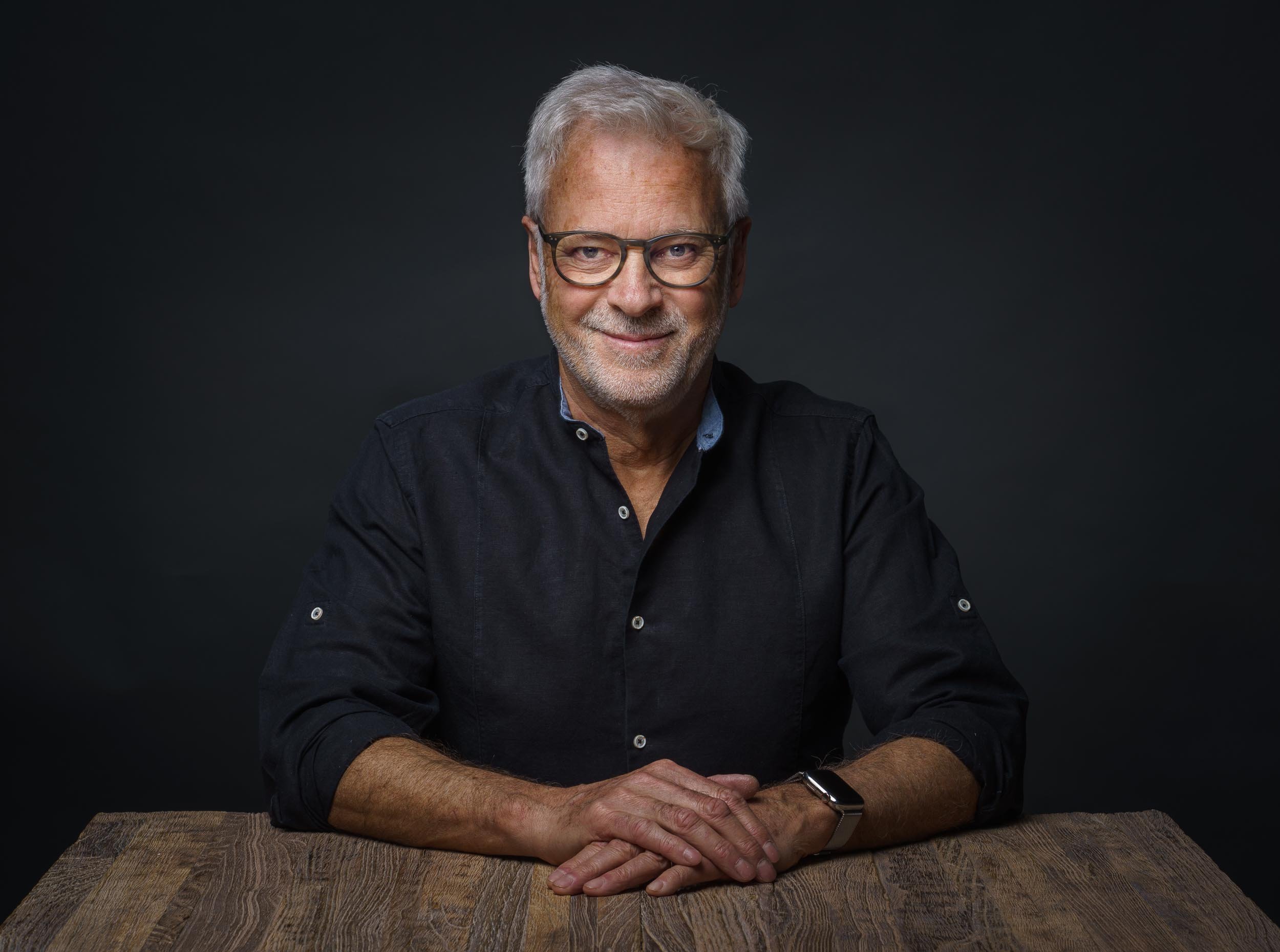 Ein älterer Mann mit Brille und grauem Haar sitzt an einem Holztisch, lächelt und trägt eine schwarze Hemd. Hintergrund dunkel.