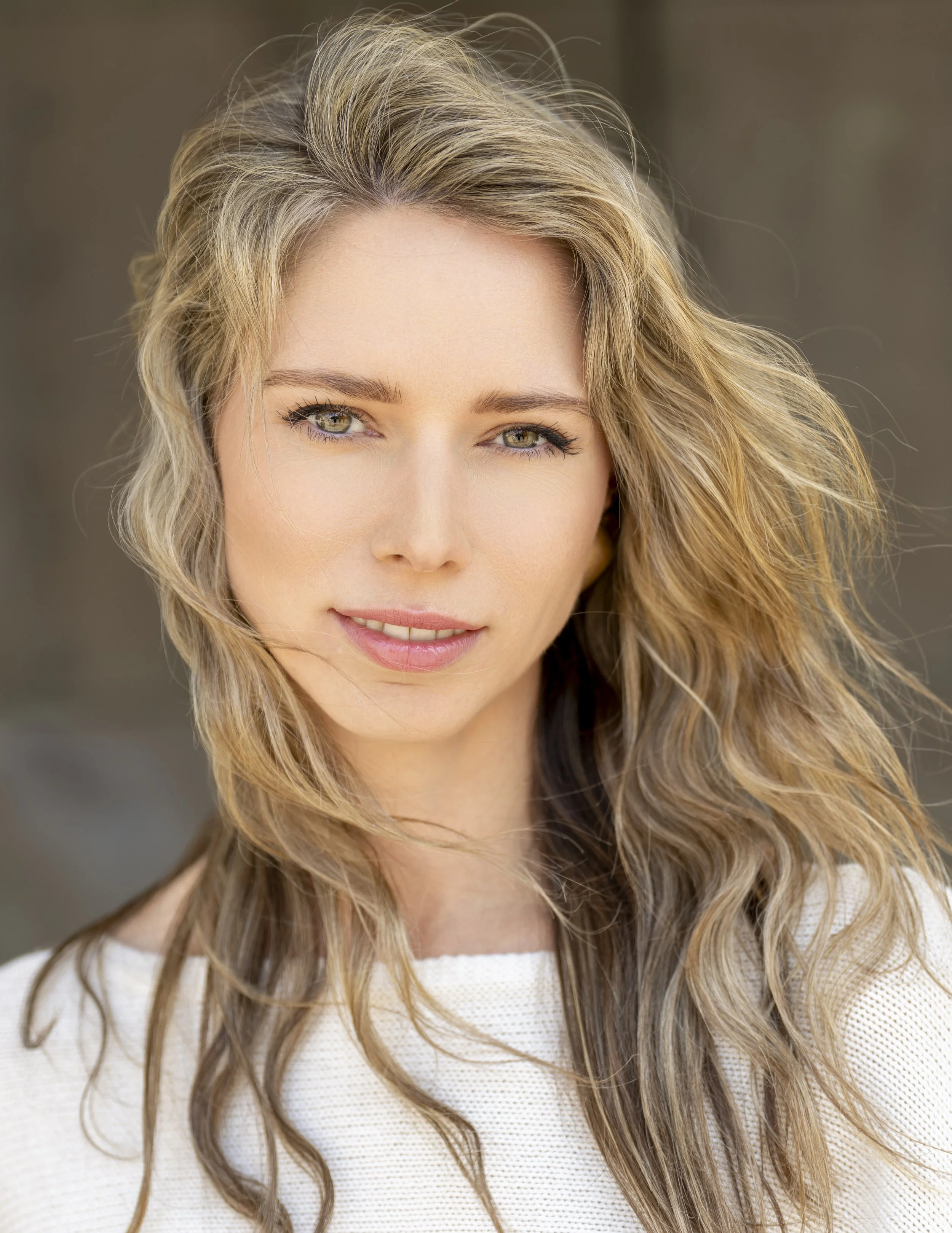 Nahaufnahme eines Frauenporträts mit lockigem, braun-blondem Haar, grün-braunen Augen und natürlichem Make-up, getragen von einer Person in einem weißen Pullover.