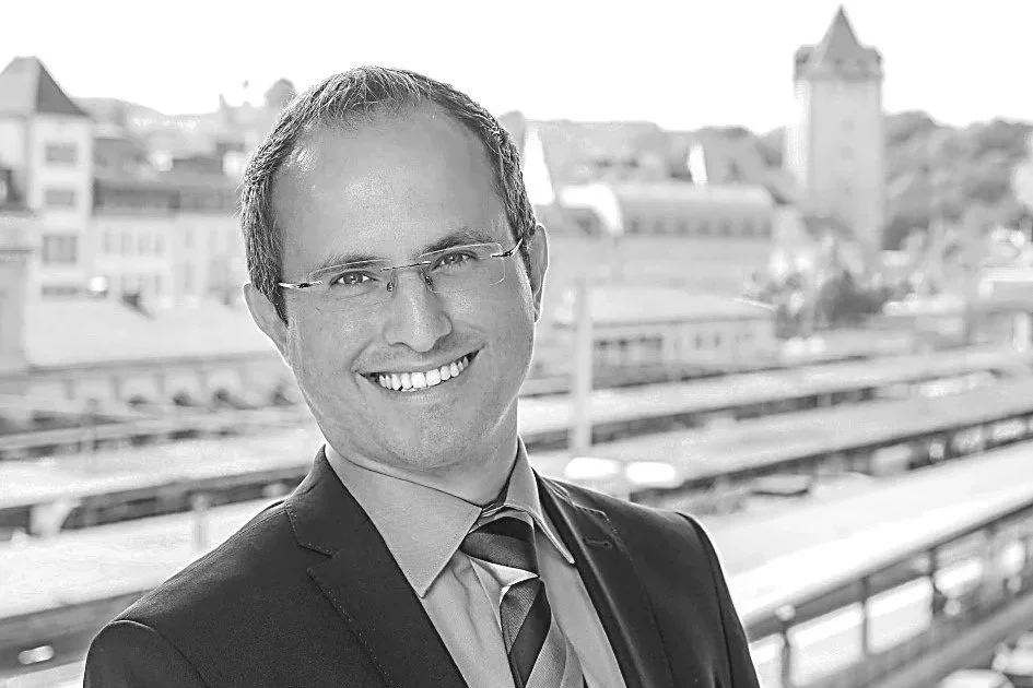 Ein Mann in formaler Kleidung, der vor einer Stadt im Hintergrund steht.