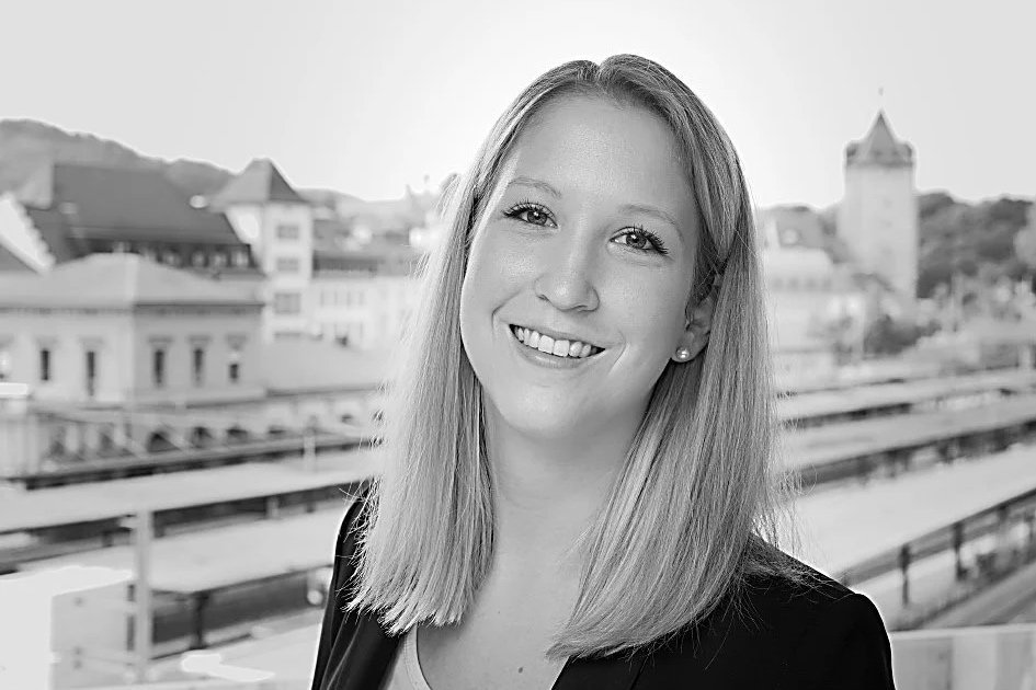 Lächelnde junge Frau mit glatten, mittellangen Haaren vor einer Stadtkulisse mit Gebäuden und einem Turm.