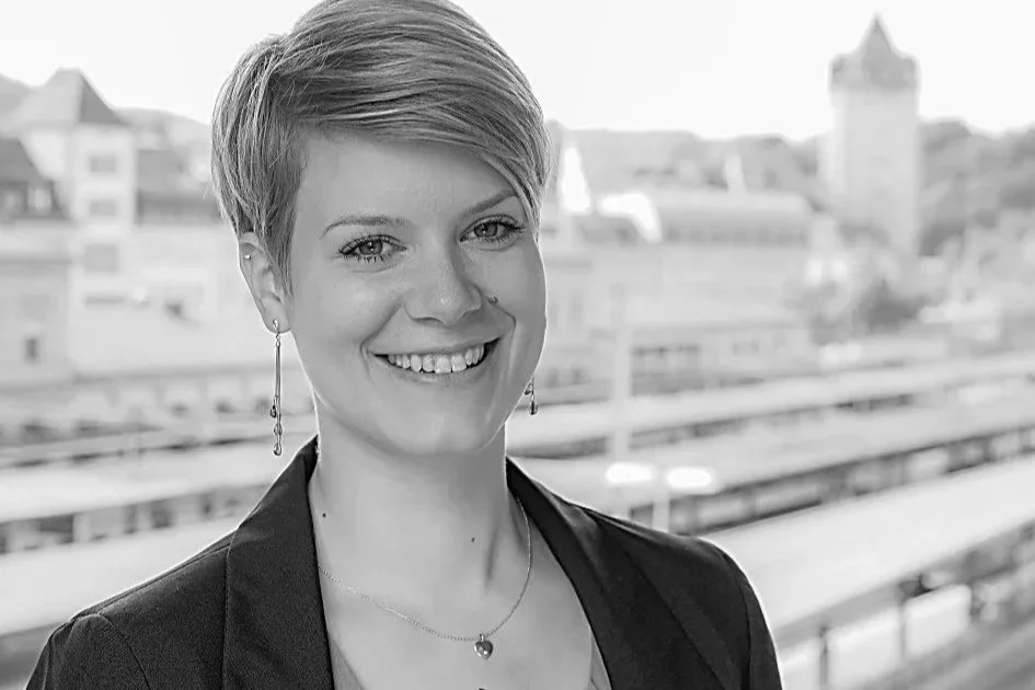 Eine Frau mit kurzem Haar, die lächelnd in die Kamera schaut, im Hintergrund eine Stadtansicht mit Gebäuden und Straßen.