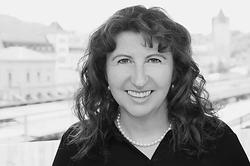 Porträt einer lächelnden Frau mit dunklem, lockigem Haar, Perlkeil und schwarzem Oberteil vor einer Stadtlandschaft.