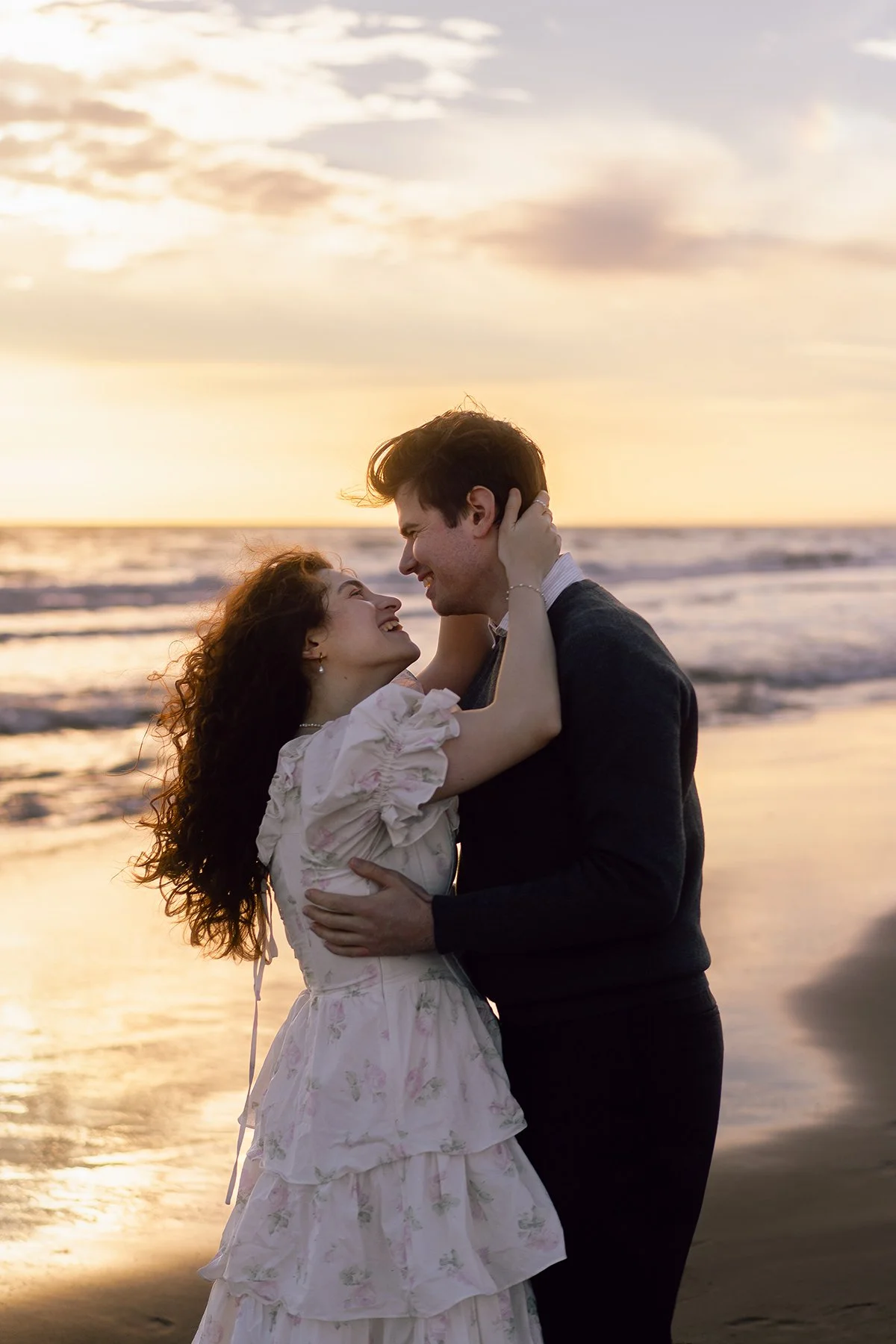 A couple embracing on the beach at sunset, looking into each other's eyes and smiling.