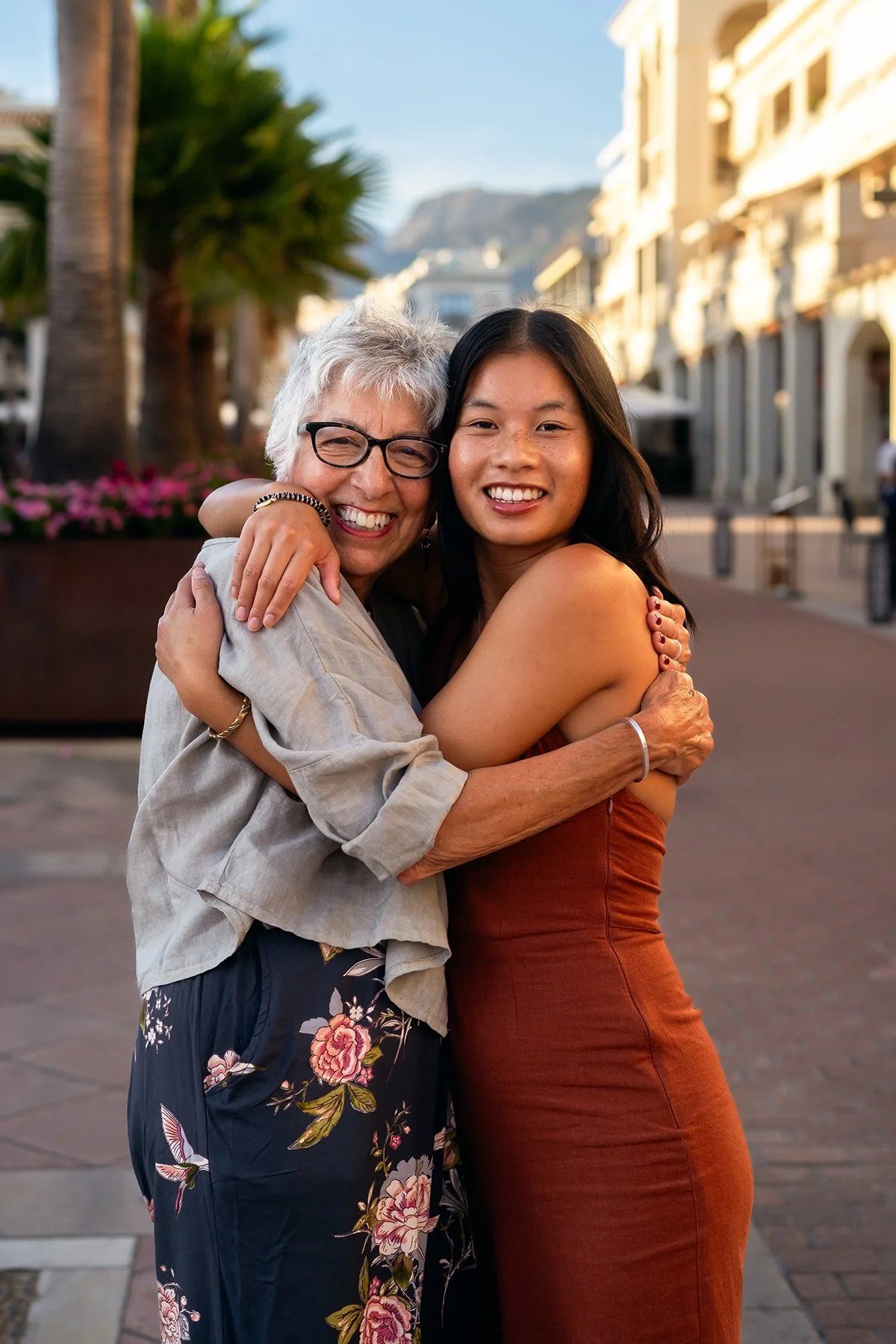 Two women hugging outdoors on a sunny day, with palm trees and buildings in the background.