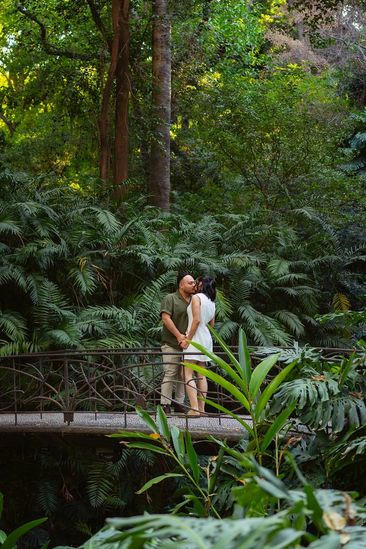 A couple sharing a kiss on a small arched bridge surrounded by lush greenery in a dense jungle.
