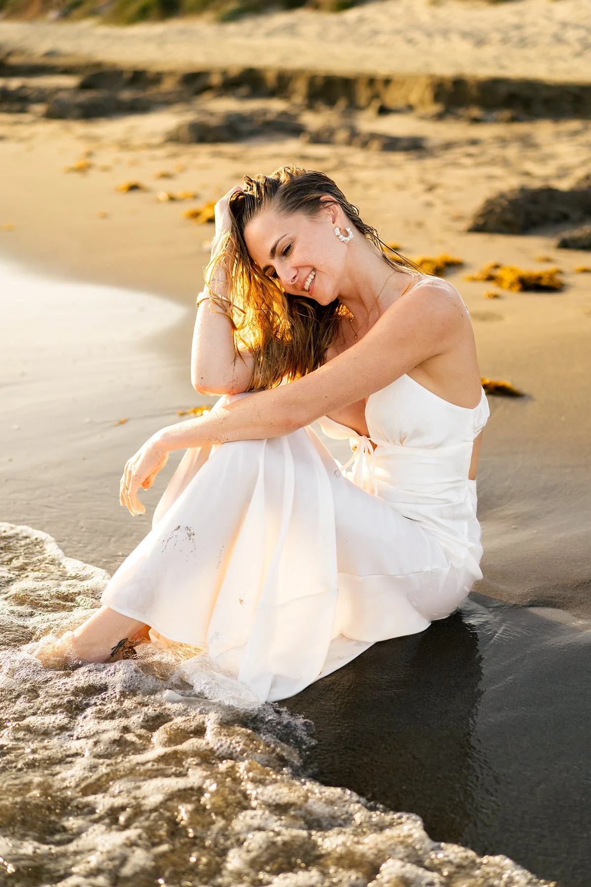 A woman with long hair and white dress sitting on the beach with her feet in the water, smiling and enjoying the sunset.