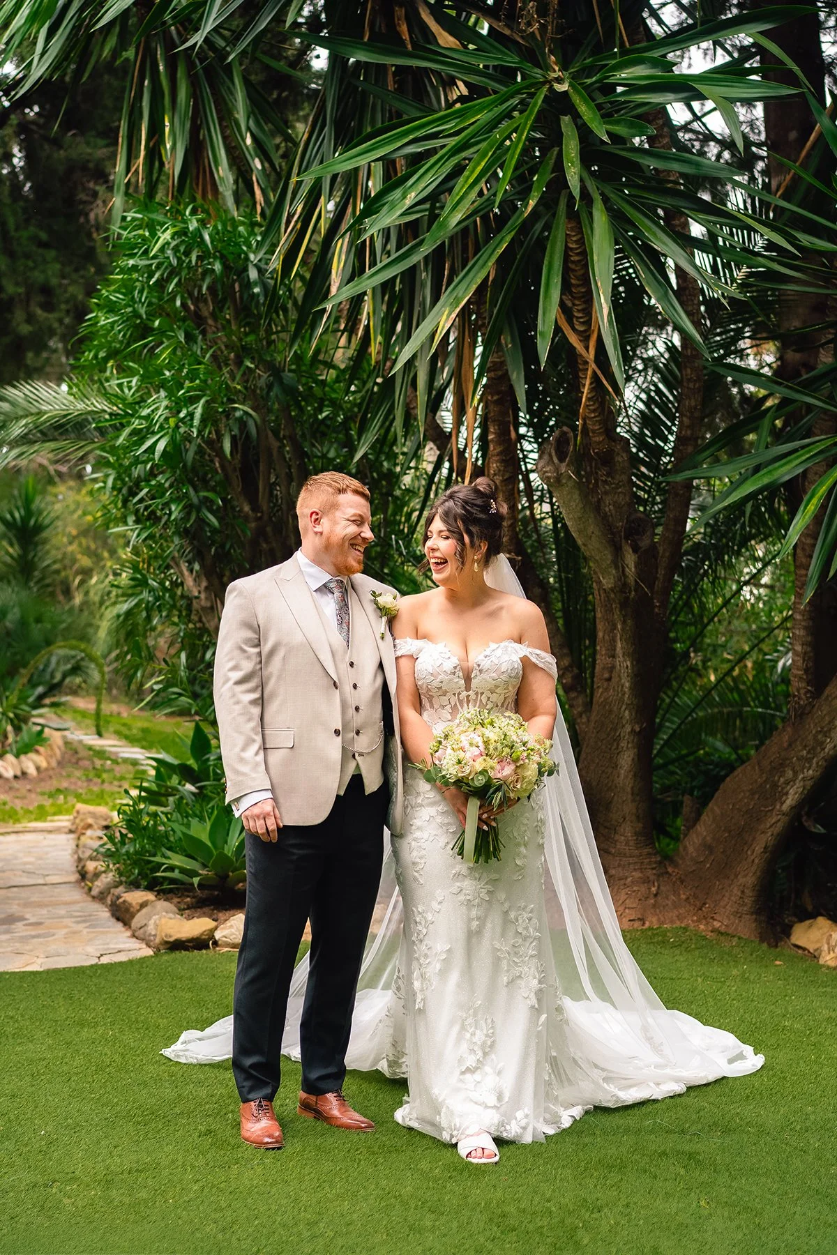 A happy couple embracing, with the woman wearing a white dress and the man in a dark suit. They are smiling outdoors, with trees in the background, suggesting a wedding or celebratory event.