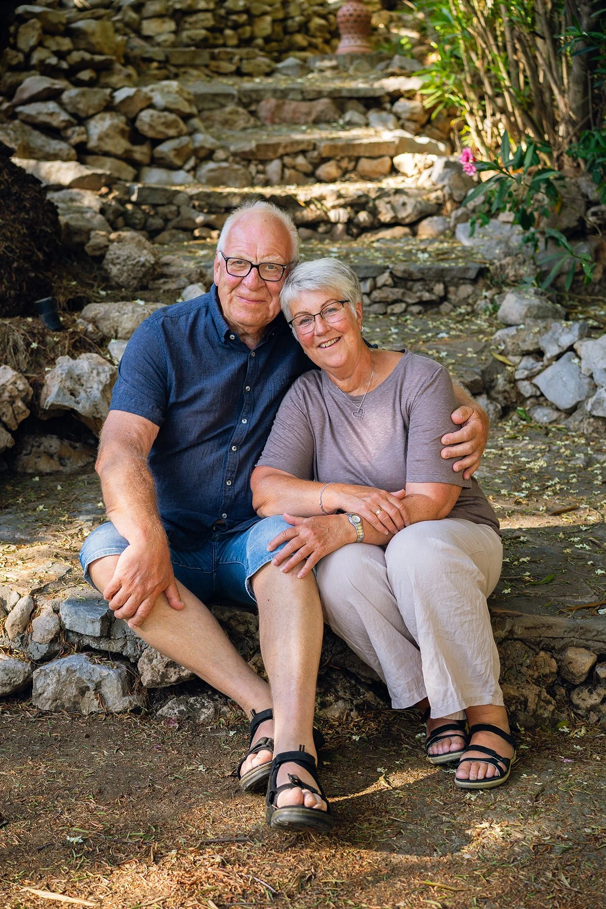 An elderly couple sitting on a stone bench outdoors, smiling and embracing, with a stone staircase and green foliage in the background.