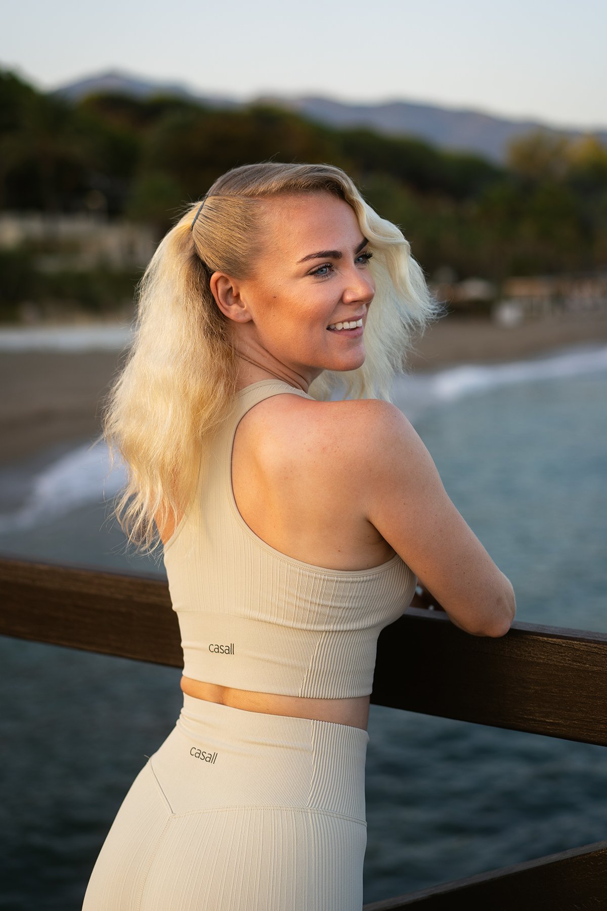 A smiling woman with blonde wavy hair and light skin leaning on a wooden railing by the ocean, with trees and mountains in the background.