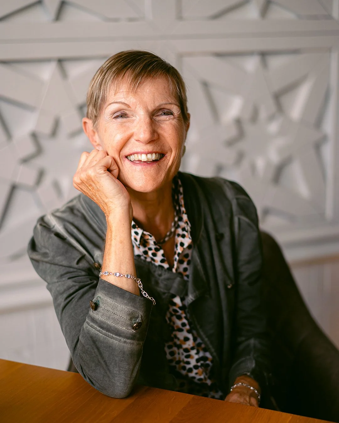 A smiling older woman with short hair, wearing a black leather jacket and leopard print blouse, sitting at a wooden table with a decorative background.