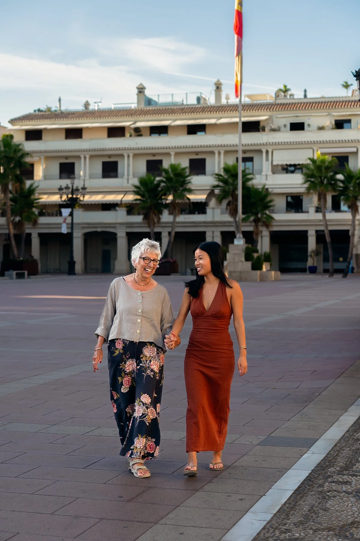 Two women walking and smiling together on a paved promenade, with a white building and palm trees in the background.