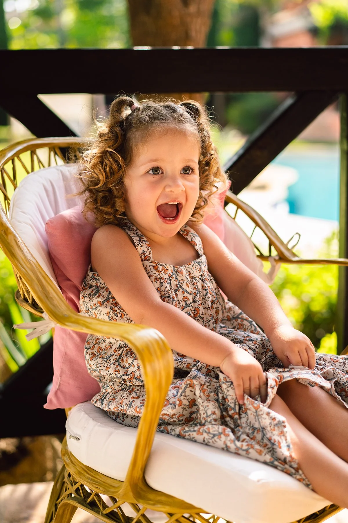 A young girl with curly hair sits in a wicker chair outdoors, with her mouth open in surprise or excitement, wearing a floral dress with a pink pillow behind her back.