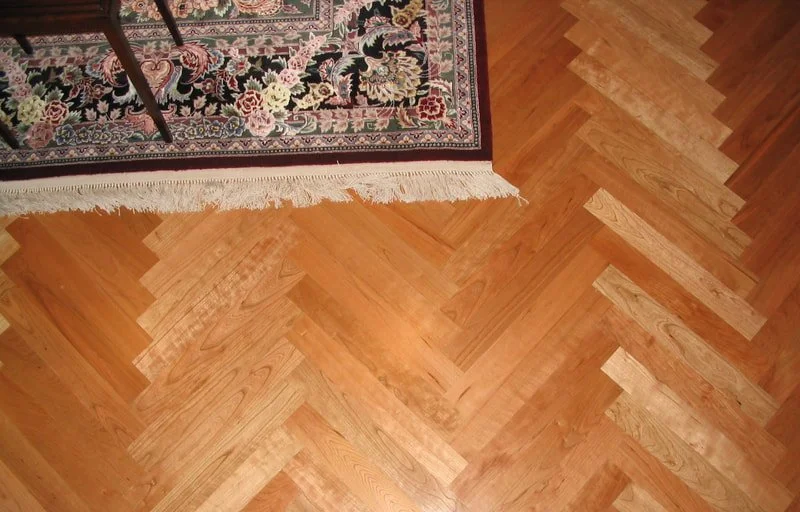 Herringbone wood floor with a fringed floral rug partially covering it.