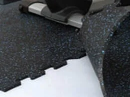 Interlocking rubber gym flooring tiles with speckled design being installed on a white surface.