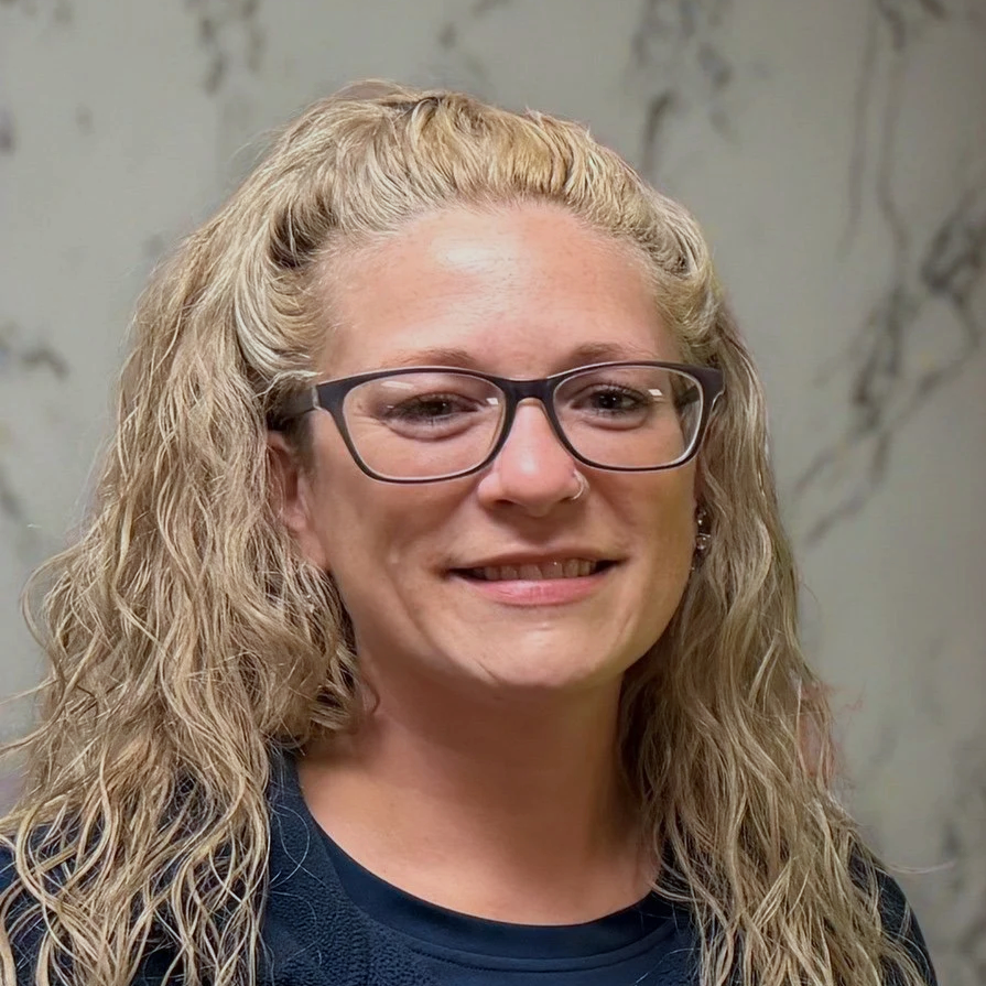 A woman with curly blonde hair, glasses, and a nose piercing, smiling in front of a marble wall.