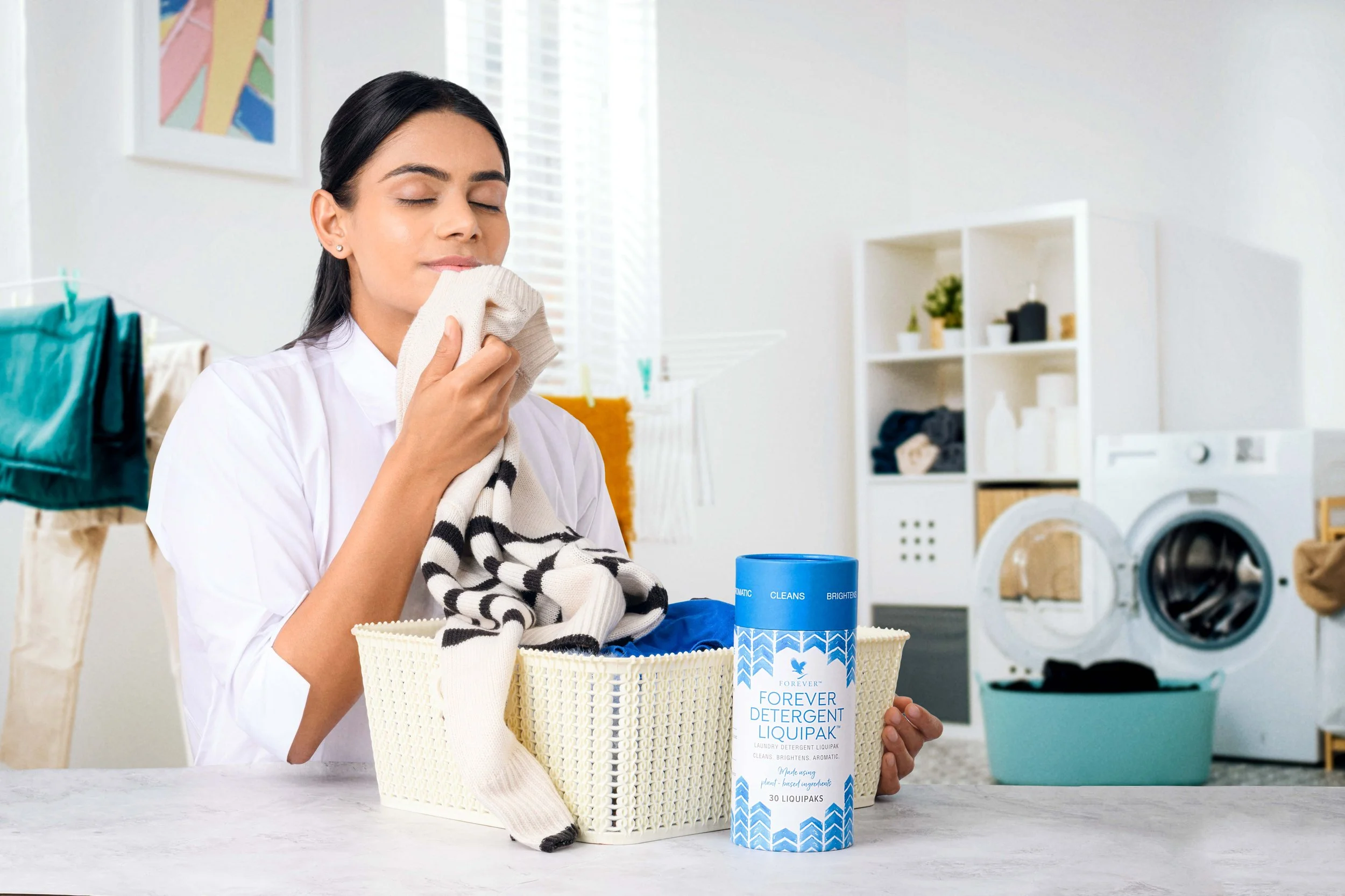 A woman with dark hair and earrings smells a laundry sock in a bright laundry room. On the table is a white basket filled with laundry, and a blue container of Forever Detergent Liquipak.