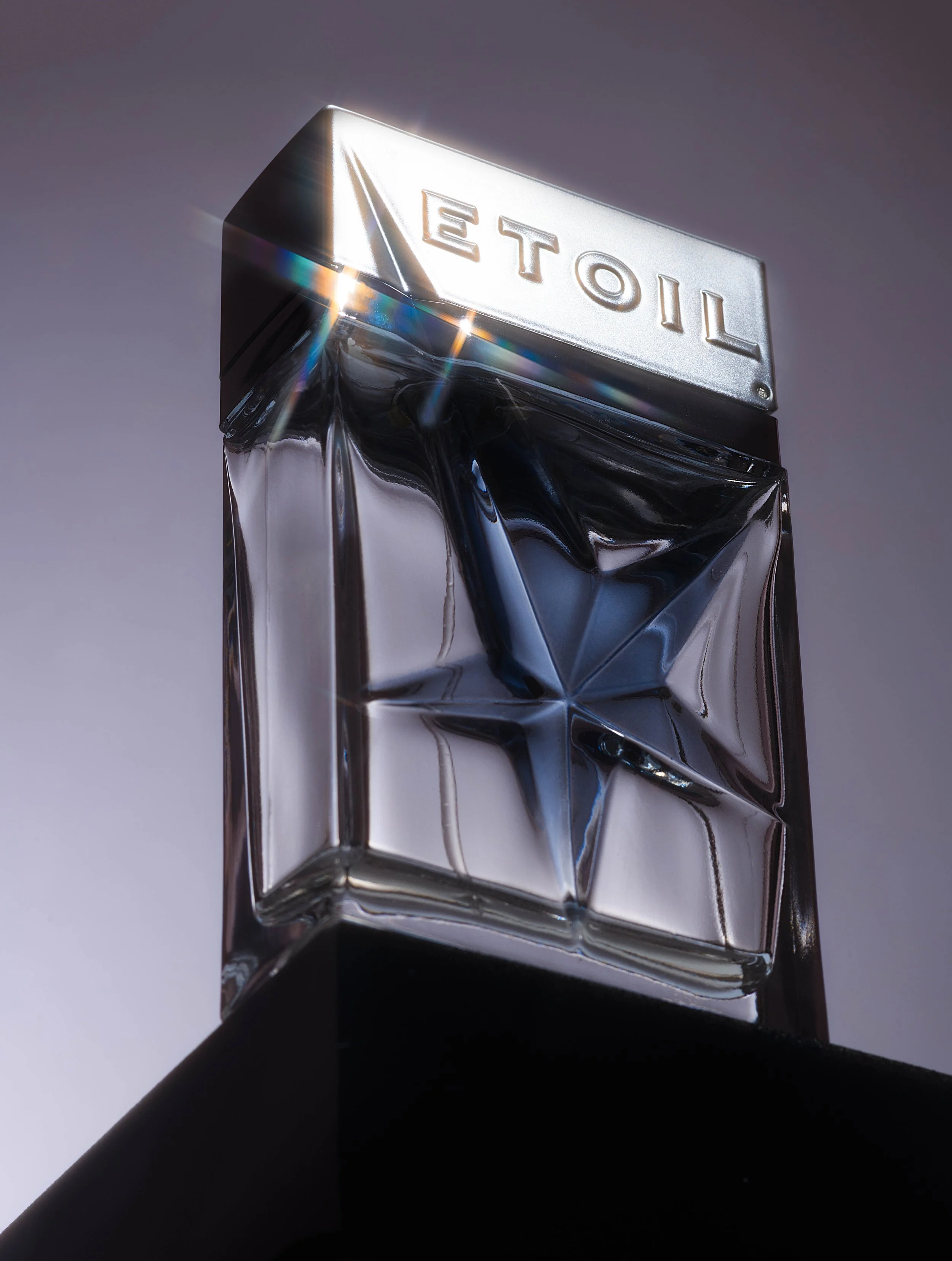 Close-up of a silver and clear glass bottle with the word 'ETAIL' embossed on the top, reflecting light and creating rainbow flares, set against a plain background.