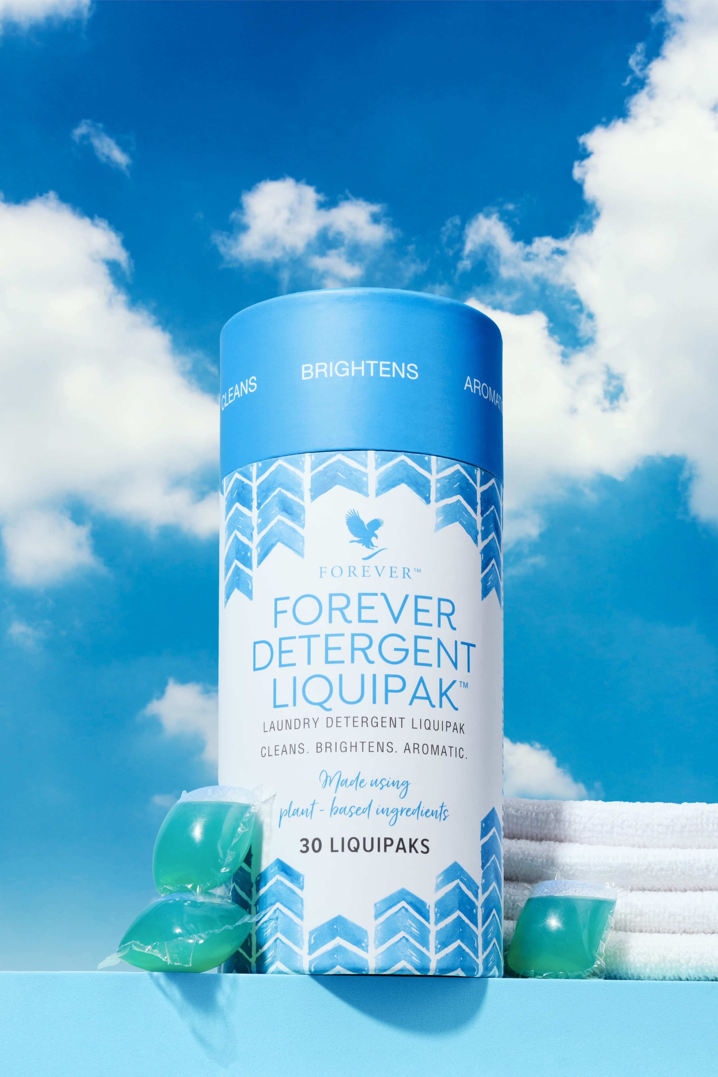 A cylindrical container of Forever Detergent LiquidPak with blue and white packaging, set against a blue sky with clouds, accompanied by folded white towels and green laundry pods.