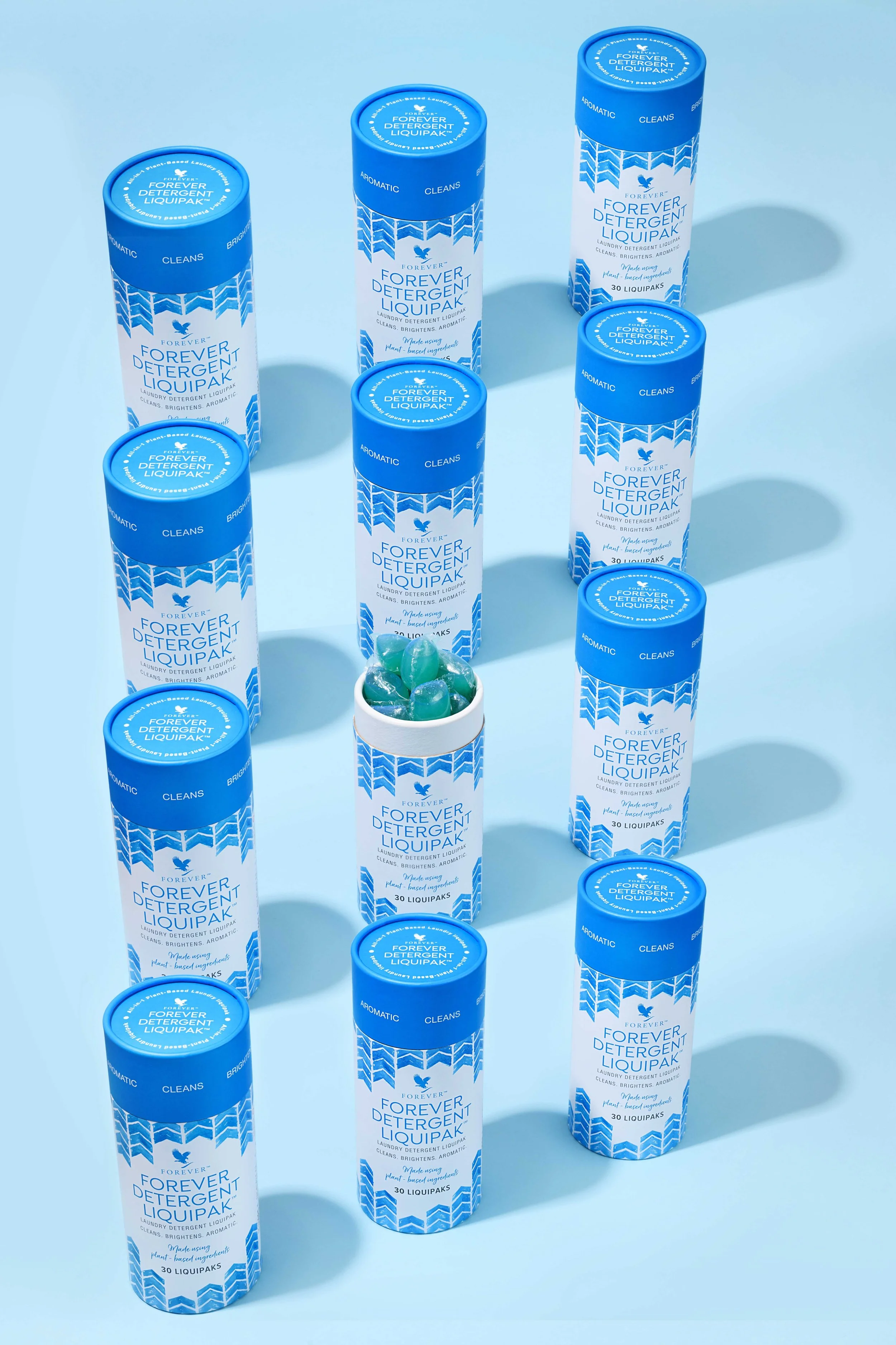 A collection of blue and white bottles of Forever Detergent Liquipak, with one open bottle showing blue detergent pods inside, arranged on a light blue background.