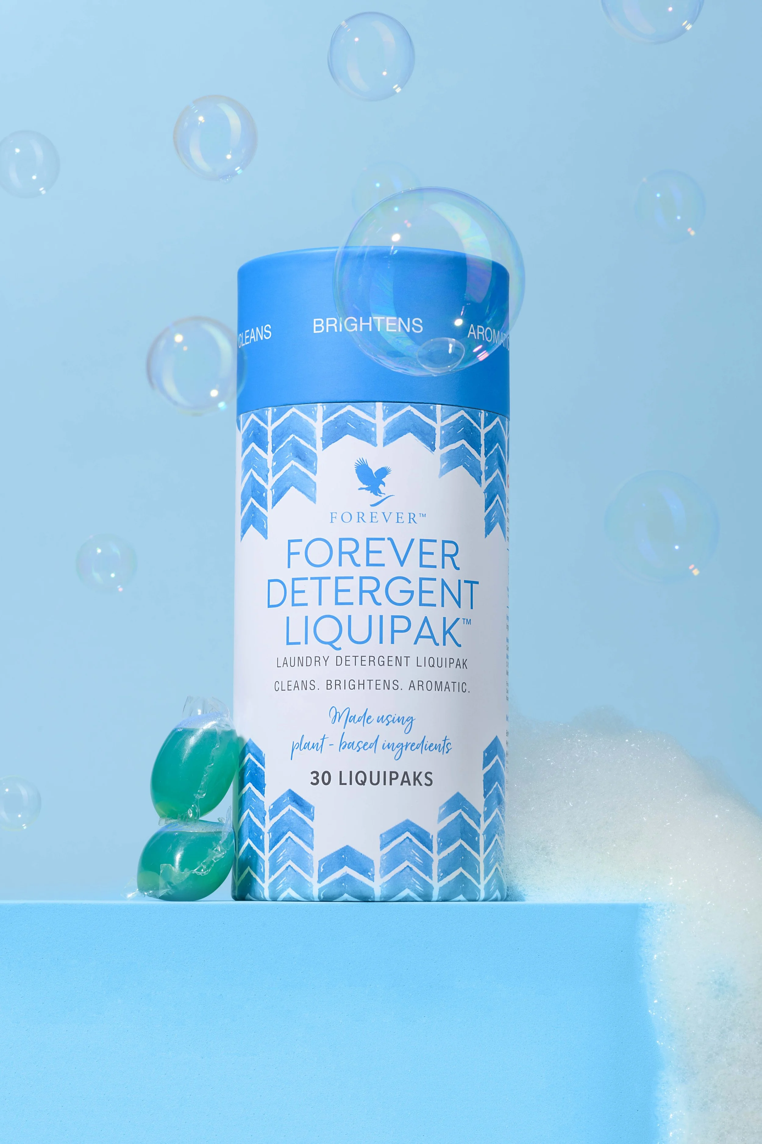 A container of Forever Detergent Liquipak with blue and white design, surrounded by bubbles and soap foam on a blue background.