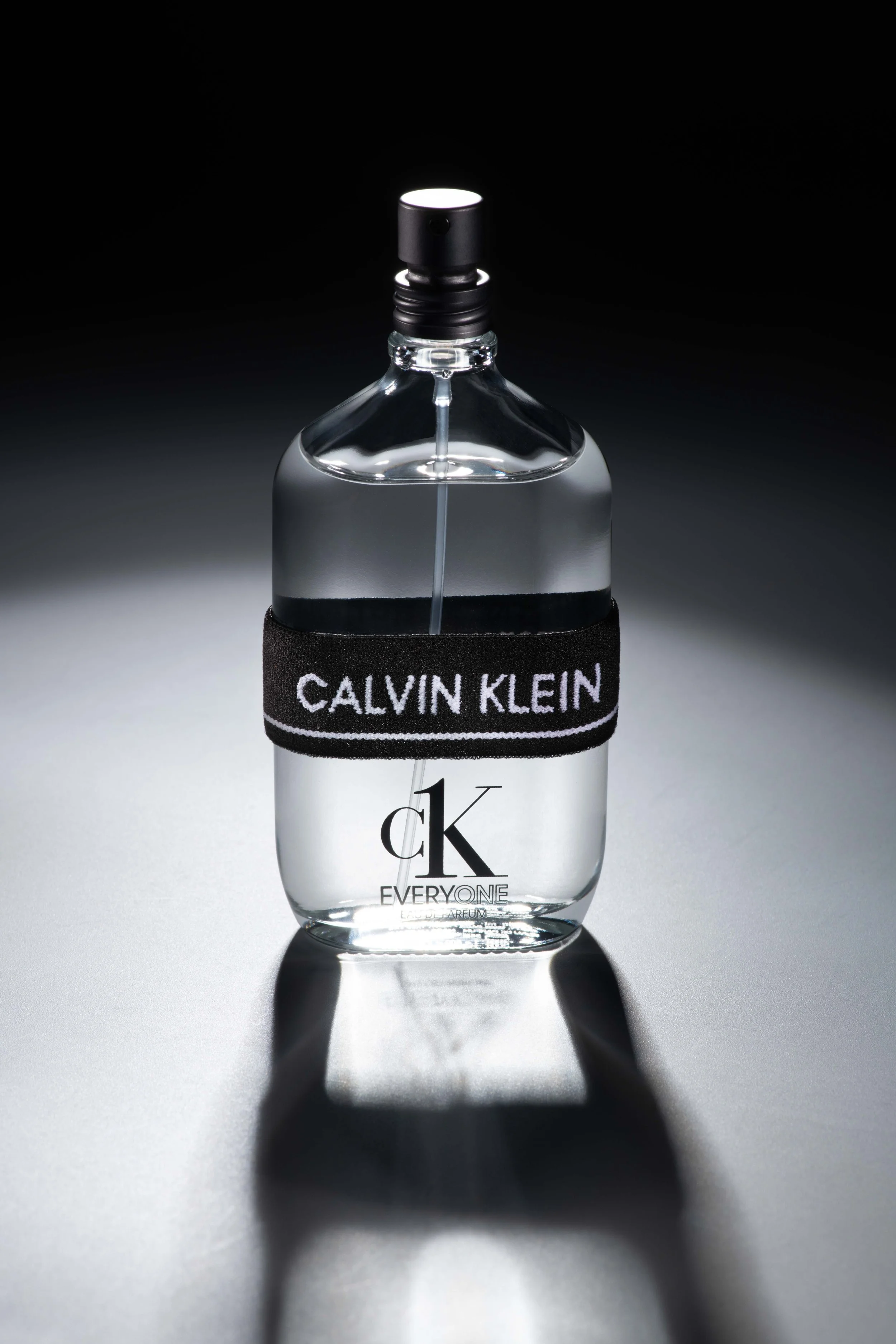 A clear glass bottle of Calvin Klein fragrance with a black band that says 'CALVIN KLEIN'. The bottle is partially filled with perfume, and a shadow is cast on the surface below.