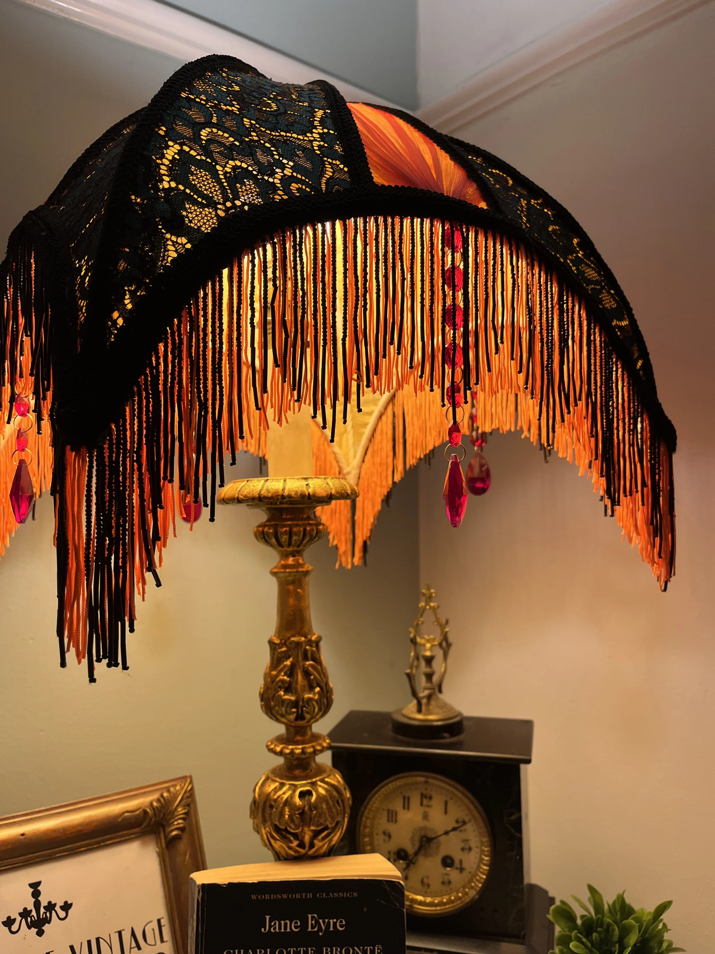 Decorative table lamp with black fringed shade, gold base, and hanging pink crystal accents. Behind it, a black vintage clock, a book titled 'Jane Eyre,' a framed picture, and a small green potted plant are visible.