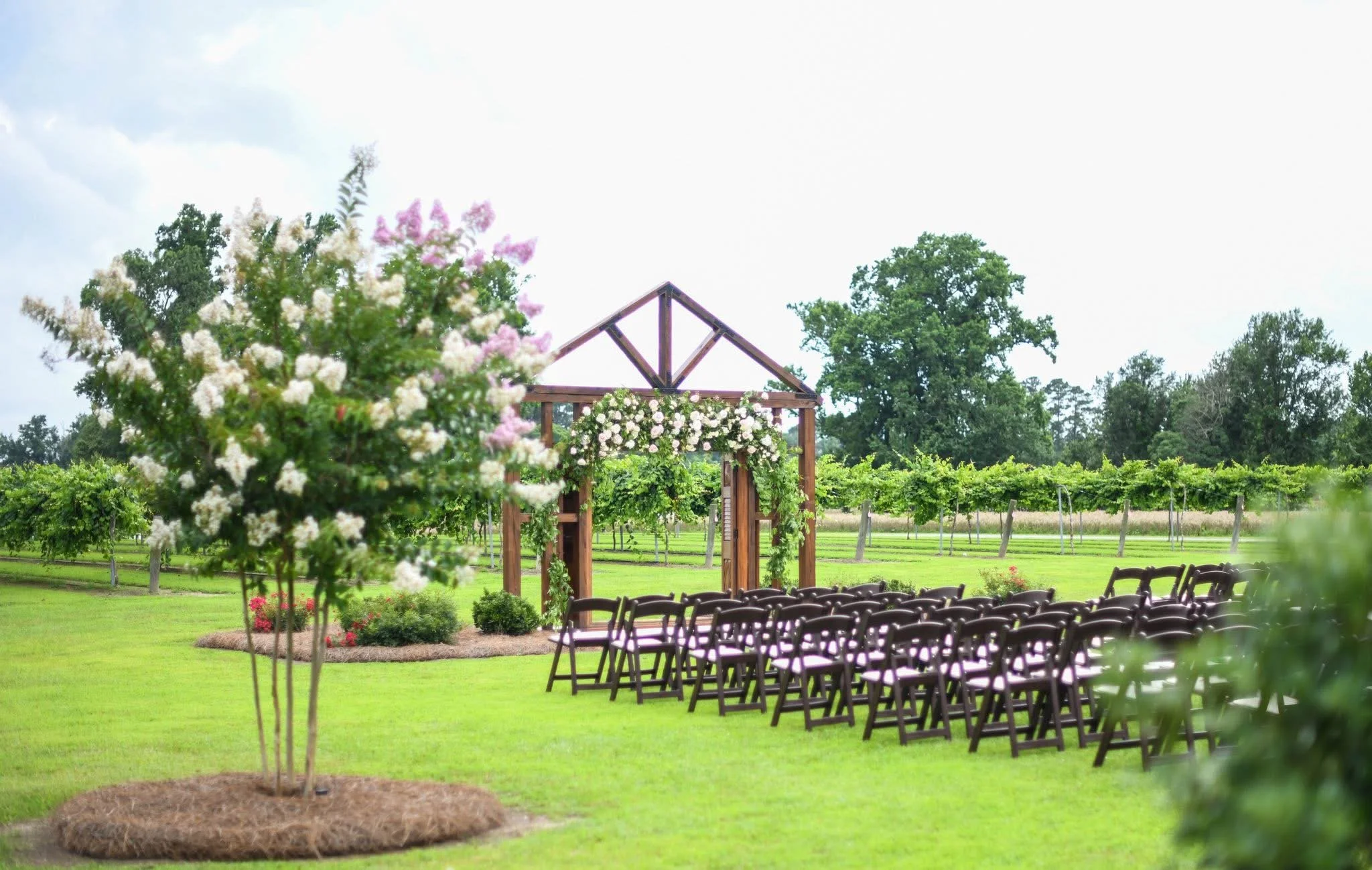 Outdoor wedding setup with chairs arranged in front of a wooden arch decorated with flowers, on a lush green lawn with flowering plants and trees in the background.