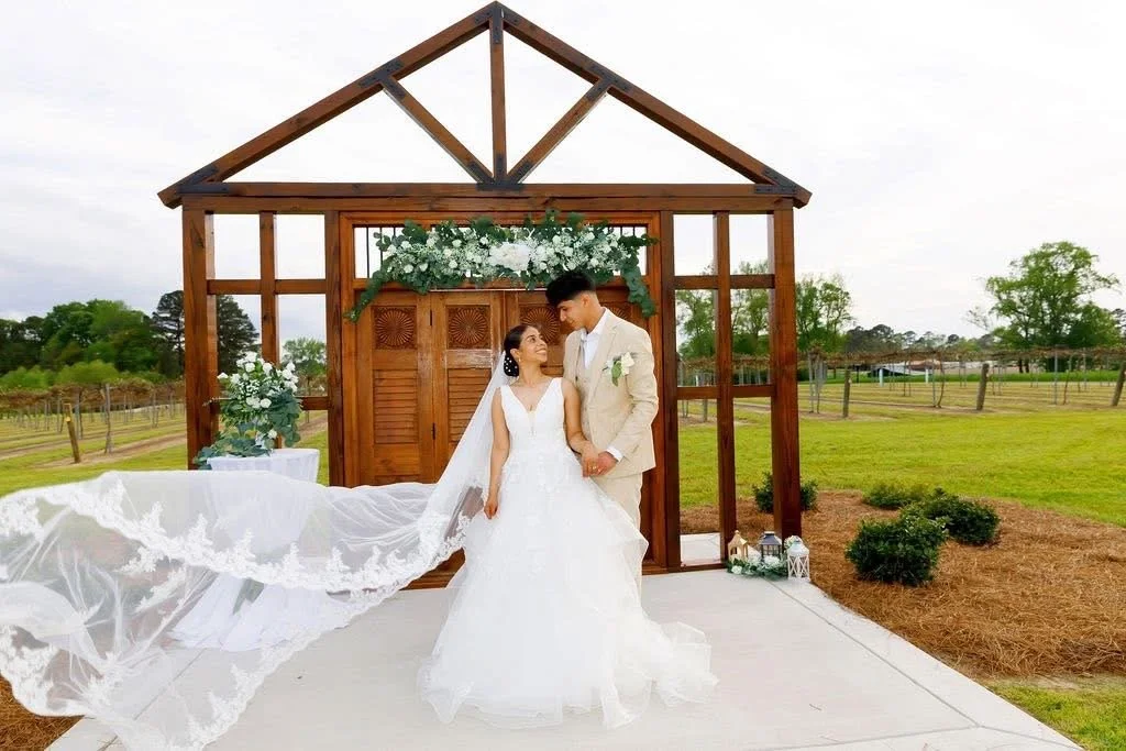A bride and groom standing together outside in front of a wooden wedding arch with white flowers, overlooking a green field, vineyard and farm scenery in Eastern NC.