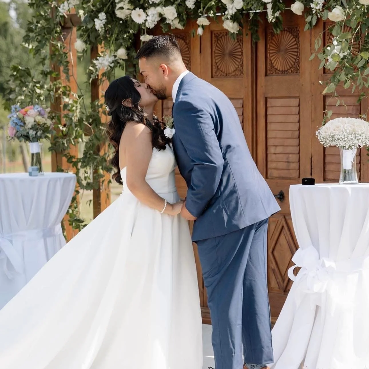 A wedding couple shares a kiss during their outdoor ceremony, standing in front of a rustic wooden backdrop decorated with flowers and greenery, at a vineyard venue in NC.