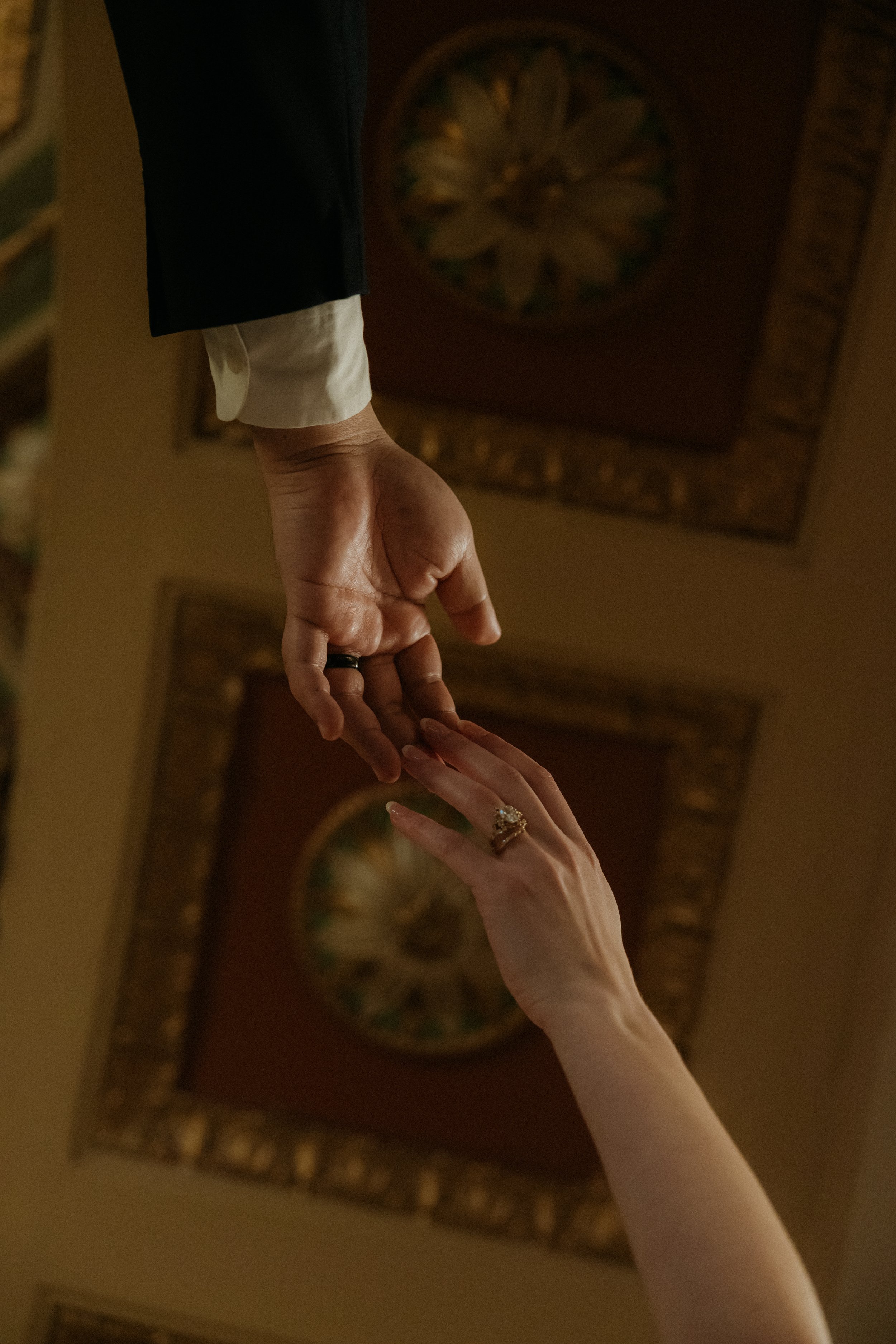 A man and woman holding hands, with the man's hand cupping the woman's fingers, both wearing rings, in an indoor setting with framed artwork on a wall in the background.