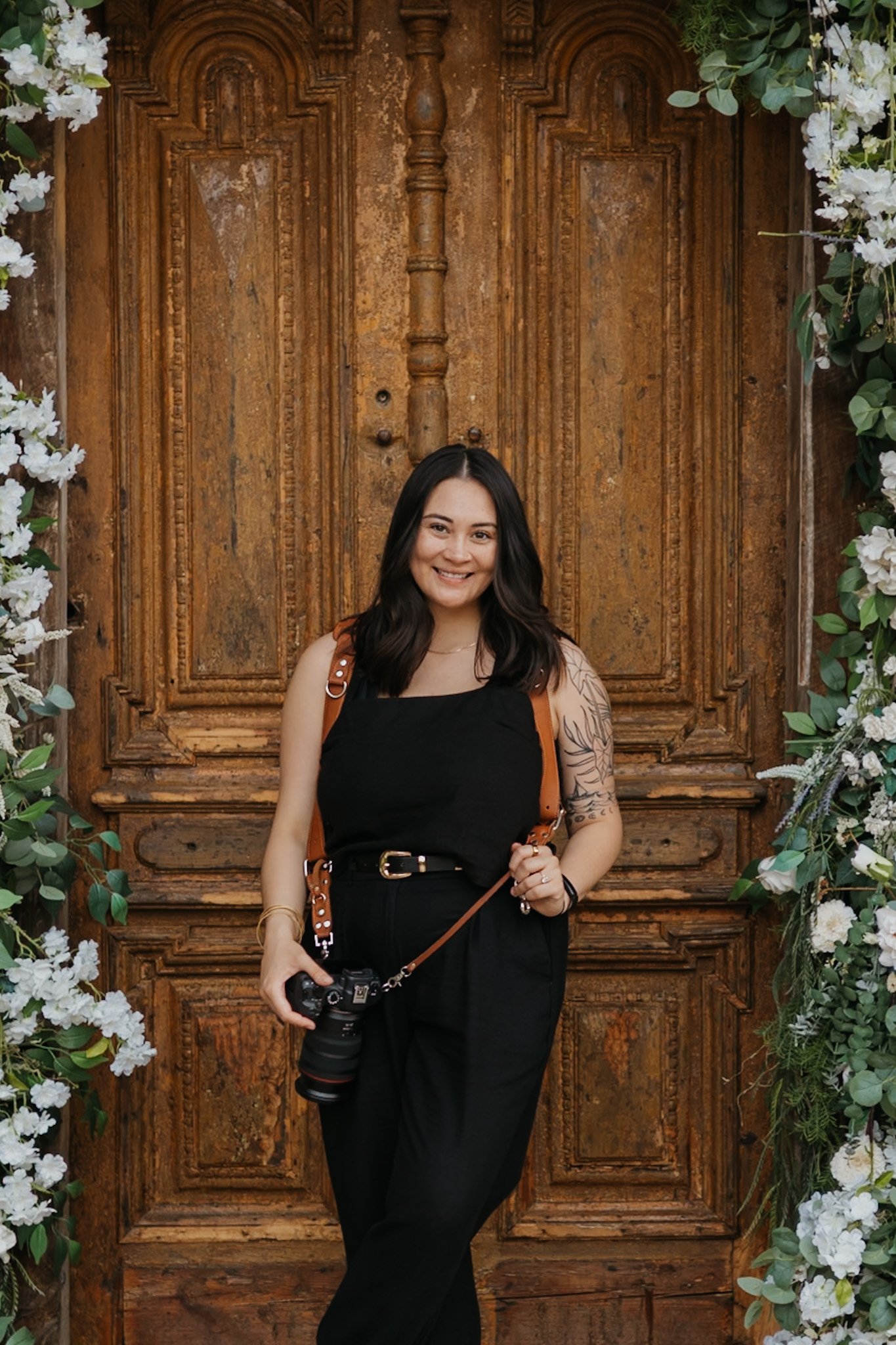 A woman with dark hair, wearing a black outfit and holding a camera, standing in front of a rustic wooden door decorated with white flowers and greenery.
