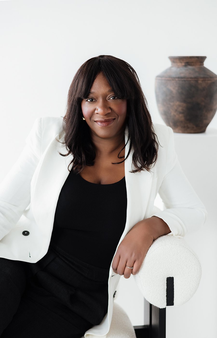 A woman with long dark hair, wearing a white blazer over a black top, sitting on a modern white chair in a minimalist room with a decorative large brown pot in the background.