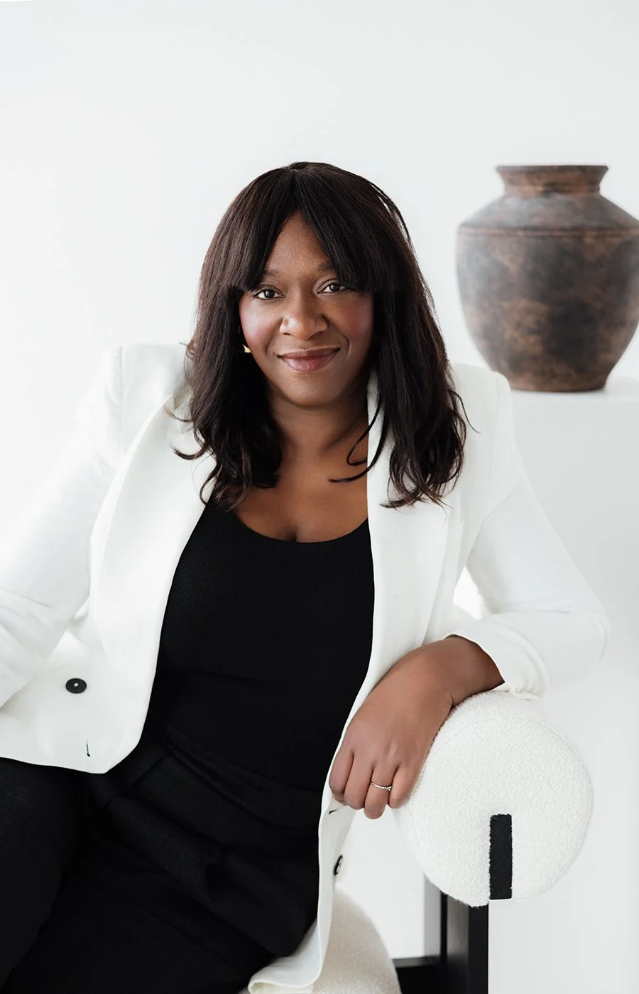 A woman sitting on a light-colored chair in a modern, minimalistic room with a white background and a large, dark brown decorative vase.