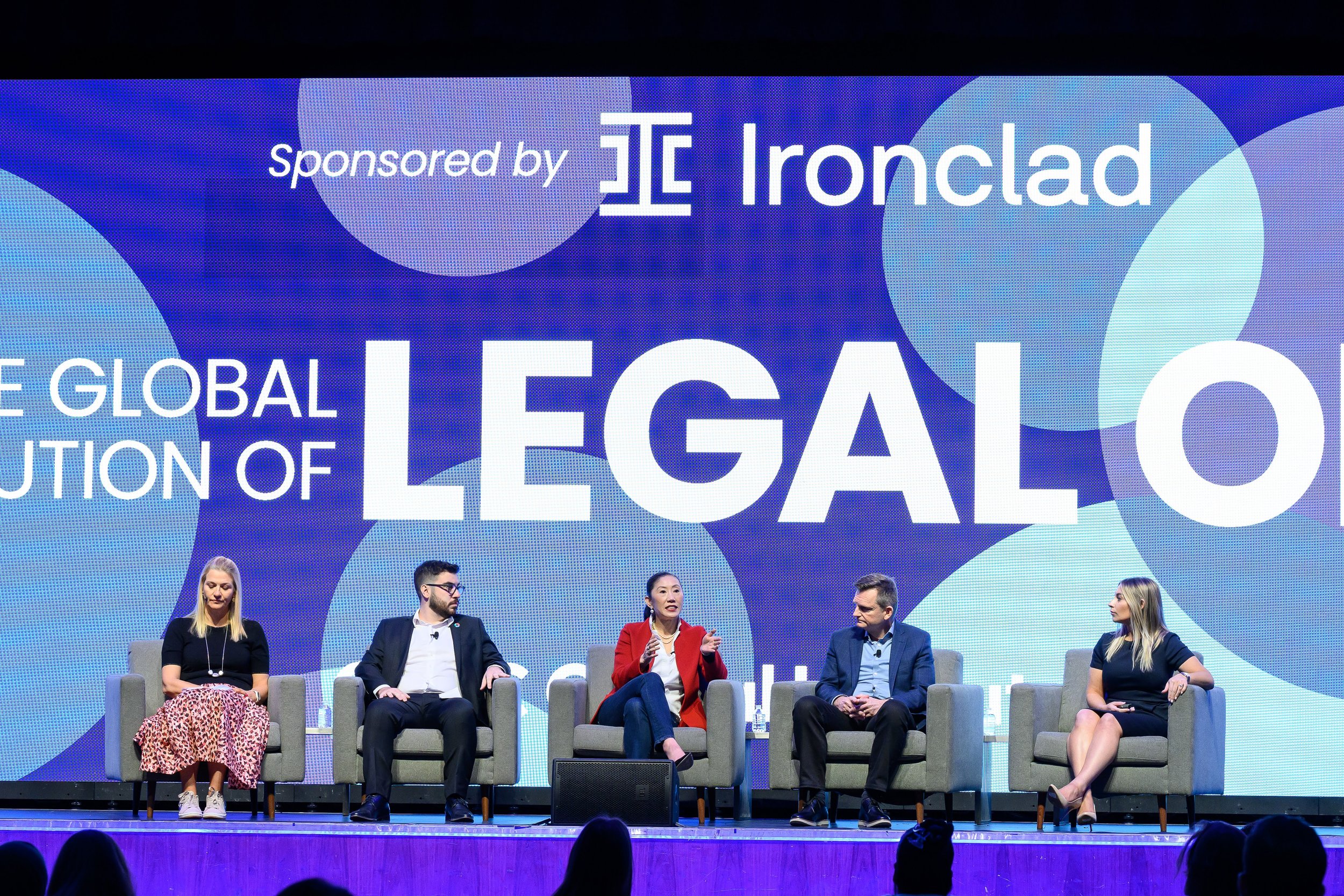 Panel discussion with five individuals seated on stage in front of a large screen displaying 'Sponsored by Ironclad' and 'The Global Revolution of Legal Ops.'