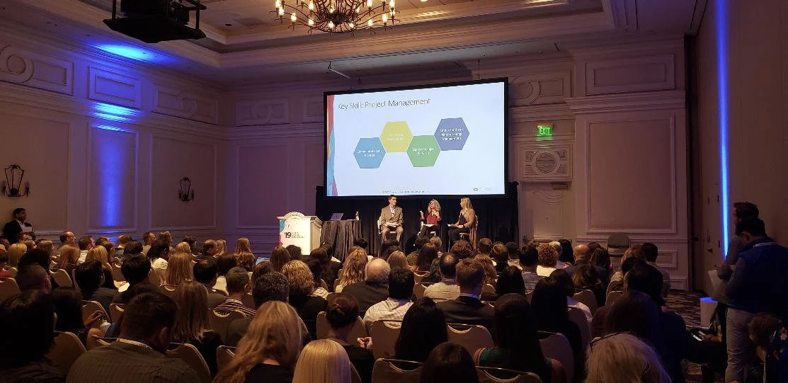 A conference room filled with attendees facing a stage where three speakers are seated. A presentation slide titled "Key Skill: Project Management" is displayed. The room is decorated with chandeliers and has a formal, professional setting.
