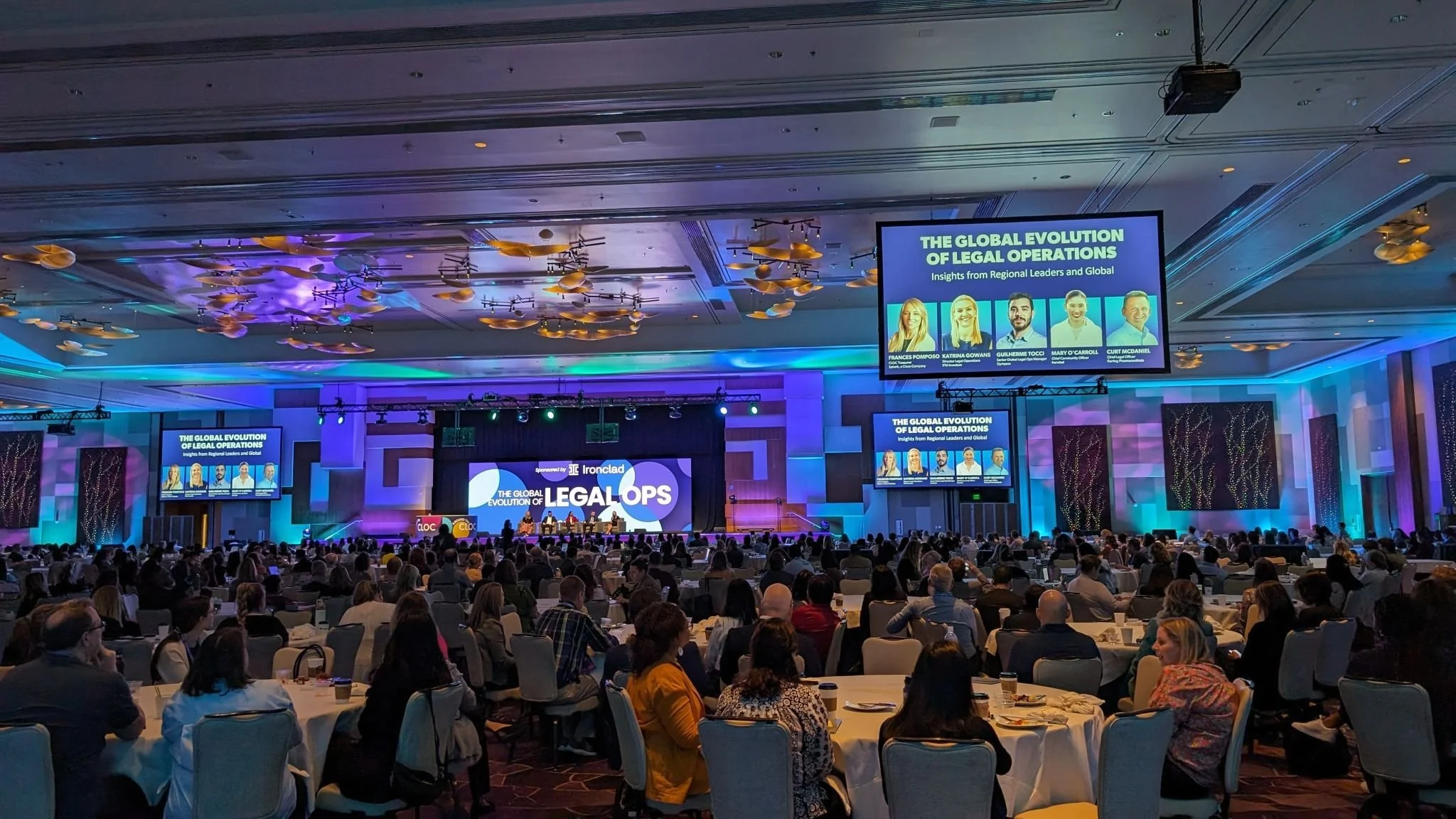 Conference room with attendees and presentation on global legal operations, featuring speakers on stage and screens displaying event details.
