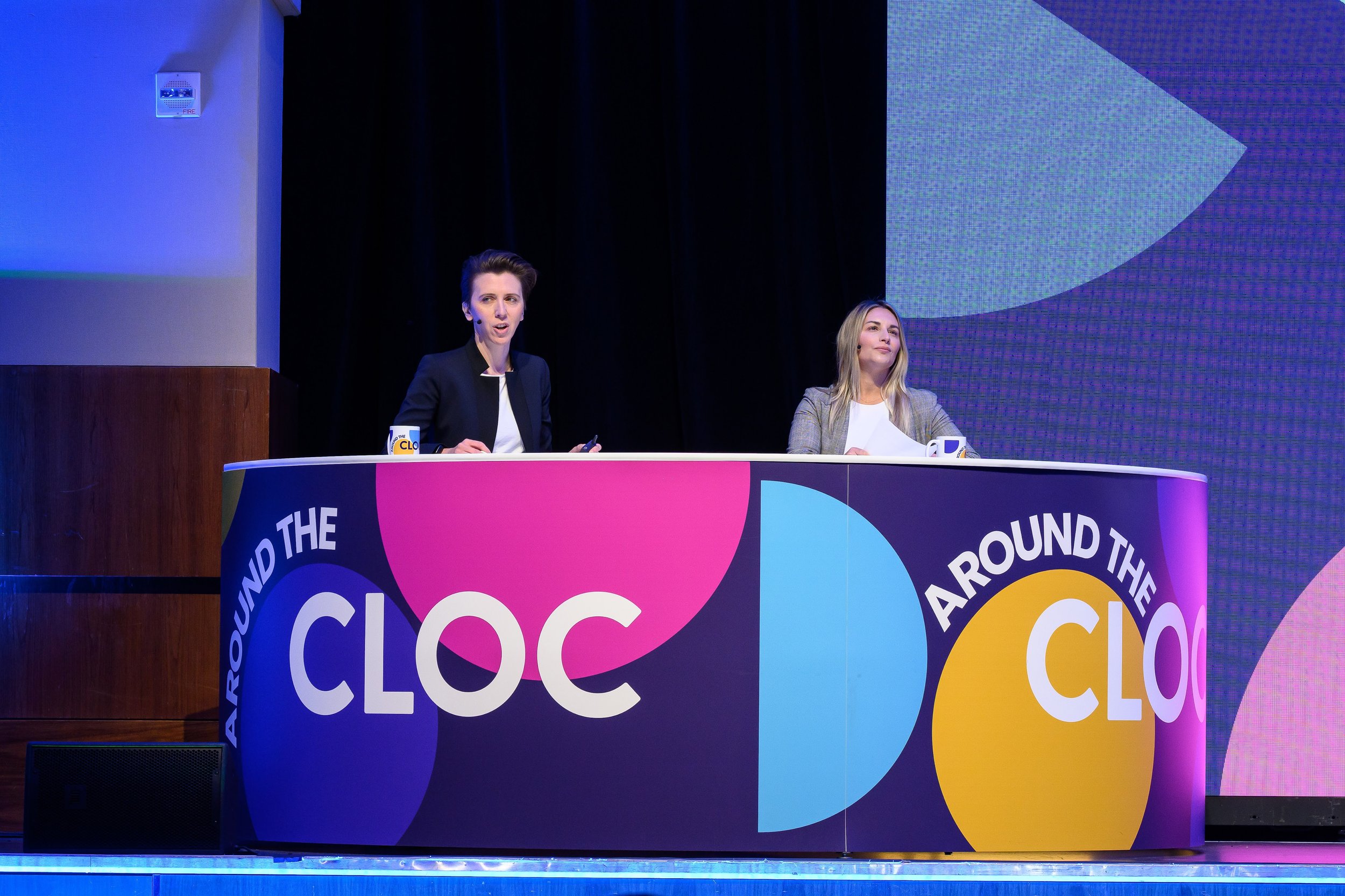 Two people sitting at a colorful desk labeled "Around the CLOC," participating in an event or conference.